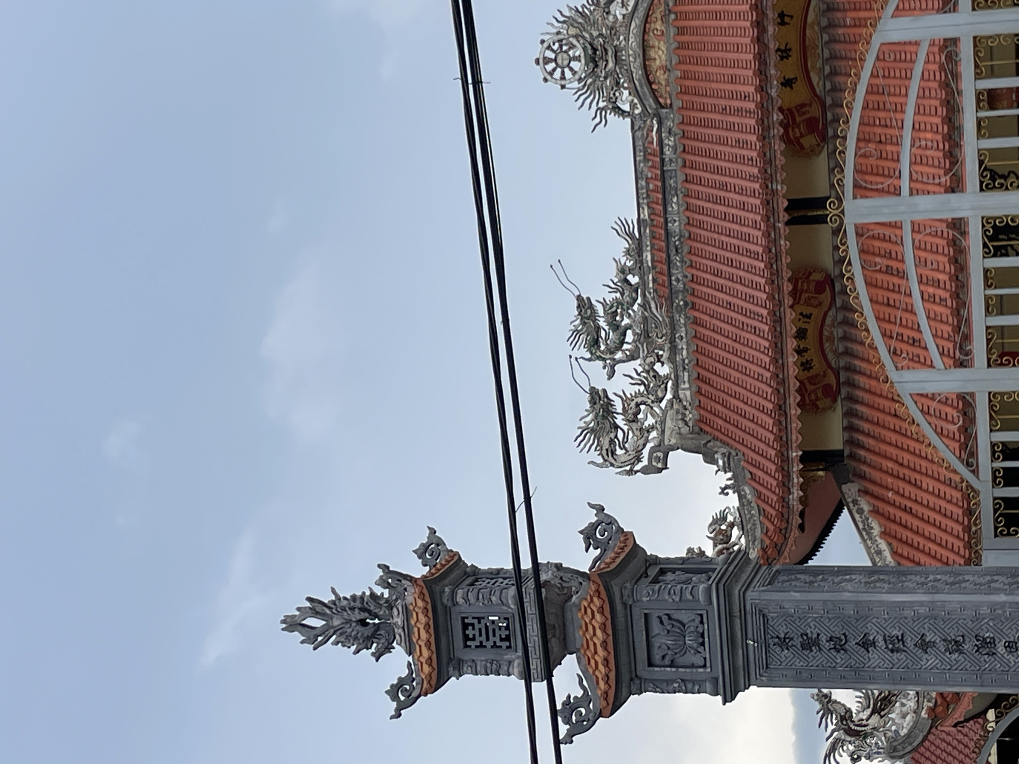 The image shows a close-up of the roof and entrance gate of a temple, likely a Buddhist temple given the decorative elements. The architecture features traditional Vietnamese design with red tiled roofs and intricate grey stone carvings. Specifically, a tall, ornate grey stone pillar with decorative carvings of lotus flowers and Chinese characters stands prominently on the left. Above the roofline, several grey dragon sculptures are visible, positioned as if guarding the temple. The roof itself is tiled with a distinctive, slightly curved design, and along the eaves, there are decorative yellow and red panels featuring more elaborate carvings. A metal gate with decorative scrollwork is visible at the bottom, partially obscuring the lower portion of the temple structure. The sky above is overcast with a pale blue hue. The presence of overhead power lines suggests an urban or semi-urban setting. While the specific location within Lâm Viên - Đà Lạt, Vietnam is not explicitly identified by signage in the image, the architectural style is consistent with temples found in this region. There are no people visible in the frame.