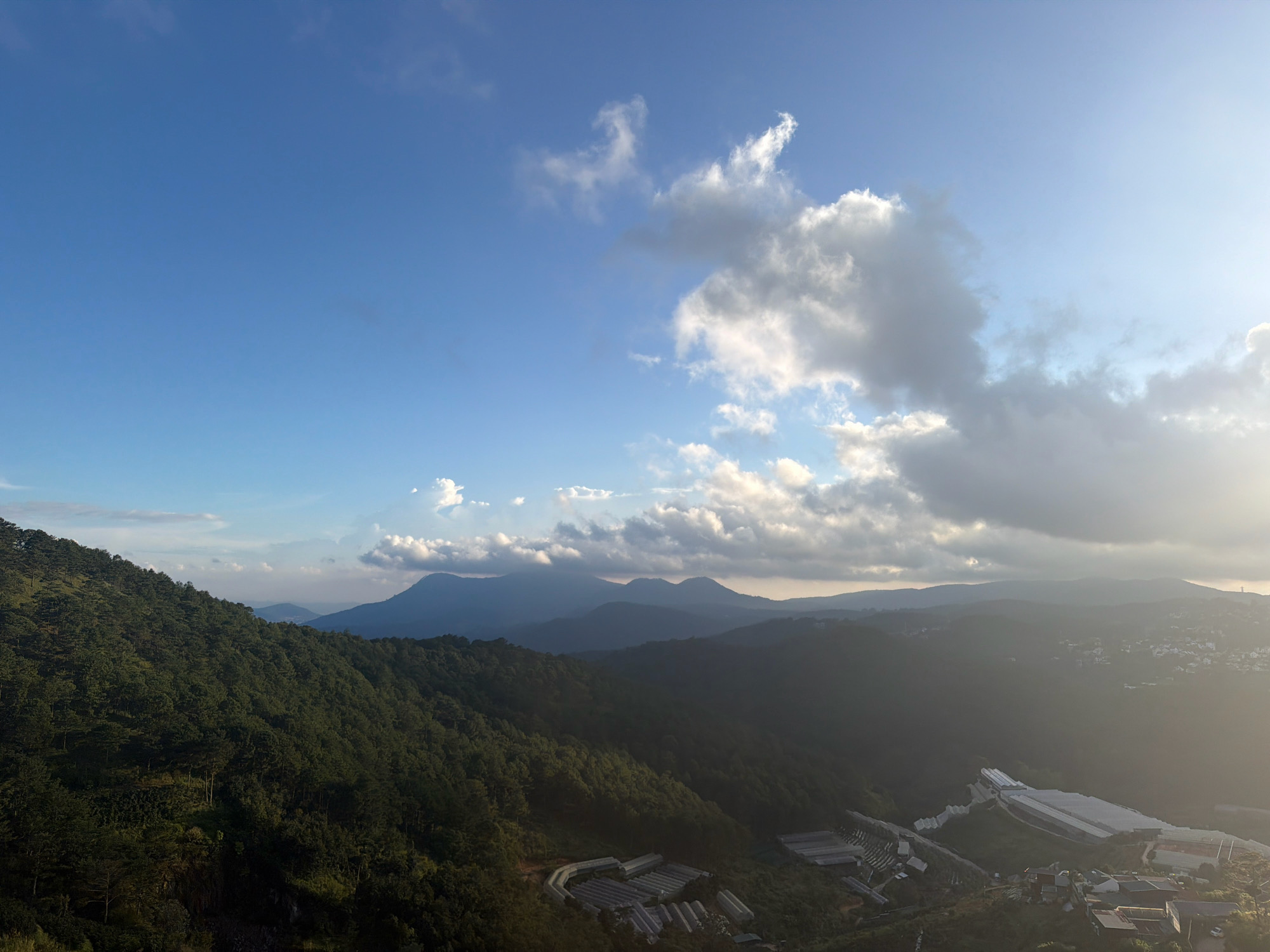 The image depicts a panoramic view of rolling, tree-covered hills under a bright, partly cloudy sky. In the foreground, a steep, densely forested hillside dominates the left side of the frame. Several greenhouses or agricultural structures with white roofs are visible in the middle ground and right, nestled within the valleys and slopes. Beyond these, successive layers of mountains recede into the distance, their forms softened by atmospheric haze. The sky is a vibrant blue with scattered, fluffy white and gray clouds, and a bright sun is visible on the far right, casting a warm glow. The scene suggests a rural or semi-rural mountainous area, possibly part of the highlands of Xuân Thành, Vietnam, known for its agricultural activities and natural beauty. No people or specific actions are discernible in the image.
