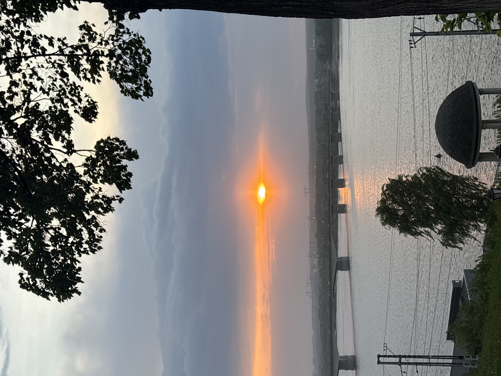 The image shows a vertical view of the Kama River in Perm, Russia, during sunset. A bridge spans the river in the mid-ground. The sun is visible in the sky, reflecting off the water surface. Vegetation, including trees, is present along the riverbank in the foreground. Power lines and structures are visible along the river. The sky is overcast with clouds.