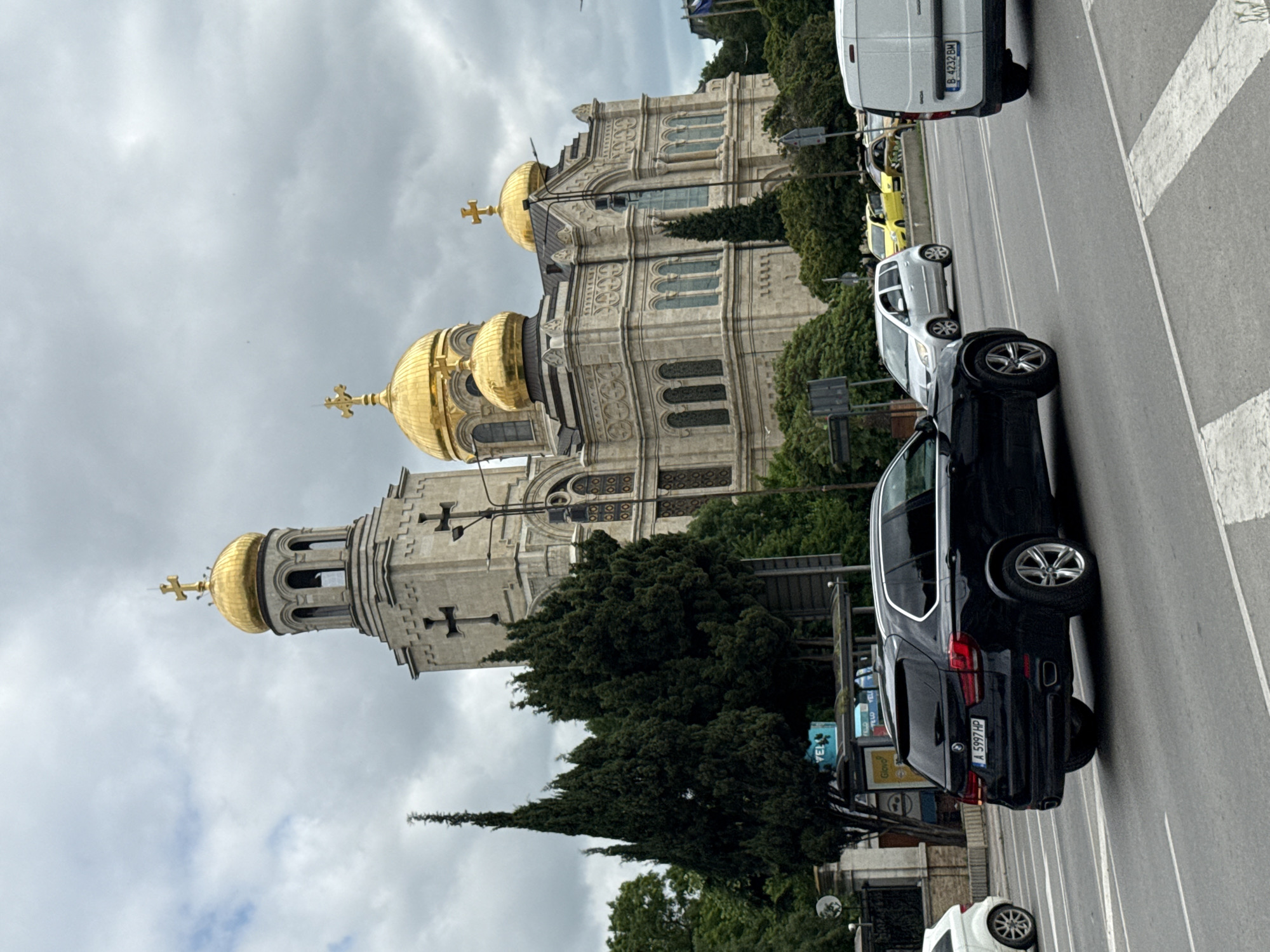 In an urban setting with a cloudy sky, a large, ornate cathedral with golden domes and crosses dominates the background. The cathedral is constructed of light-colored stone and features intricate architectural details. In the foreground, several cars are on a multi-lane road. A black BMW SUV is in the left lane, followed by a white van in the right lane. Other vehicles, including a silver sedan and a yellow car, are visible further down the road. Trees and traffic signals are interspersed throughout the scene. A bus stop shelter is present on the left side of the road, with advertisements displayed.