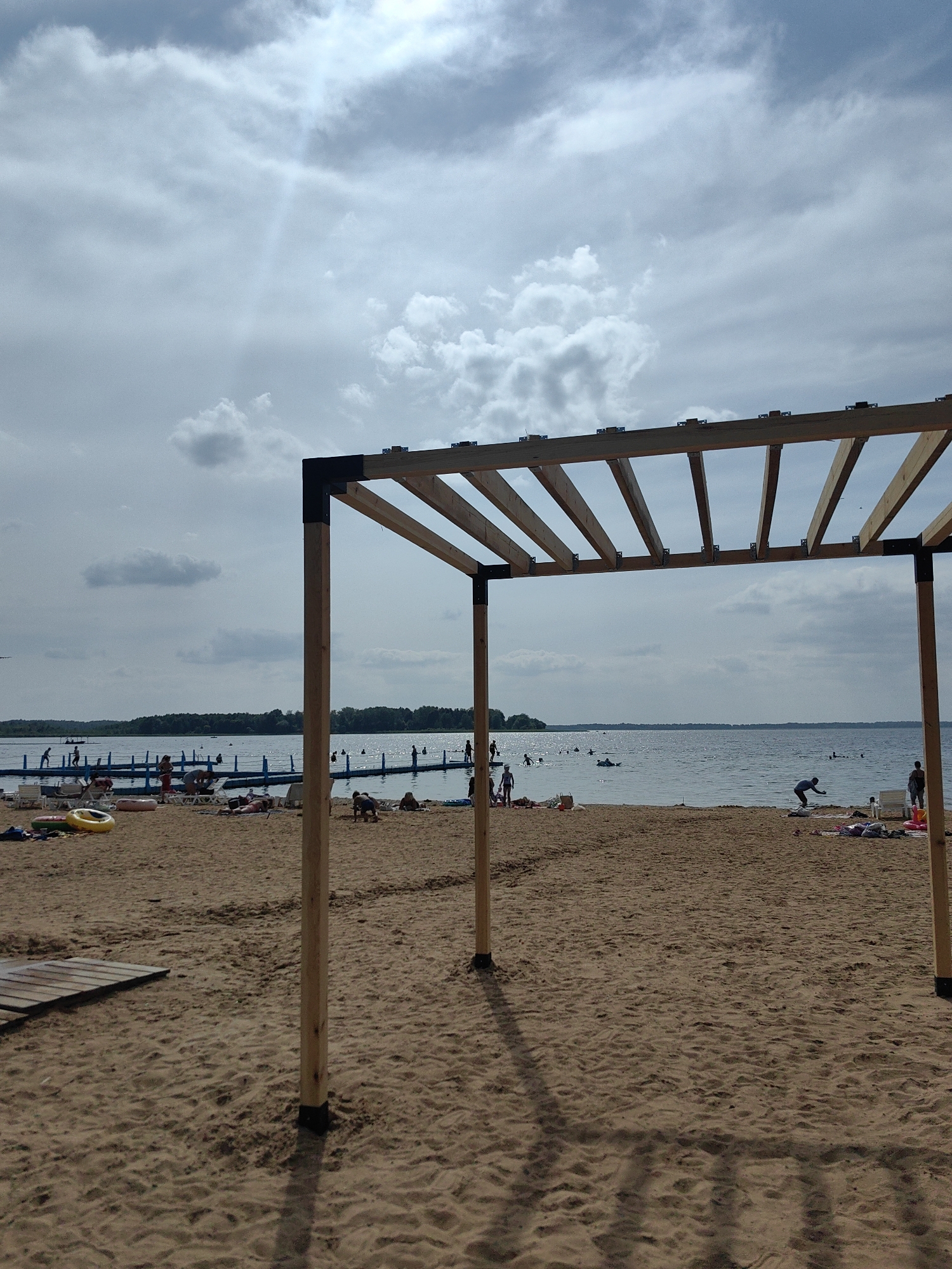 This image depicts a sandy beach scene on a cloudy day, likely at a waterfront location in Myadzyel, Belarus. In the foreground, a partially constructed wooden pergola stands, with its frame casting shadows on the sand. The beach is populated with a few individuals. To the left, there are beach chairs and a yellow inflatable ring. Further back, a blue floating barrier delineates a swimming area in the water. Several people are visible in the water, some swimming and others standing. The water is calm, and a tree line is visible in the distance on the far shore. The sky is mostly overcast with some brighter areas where sunlight is breaking through the clouds.