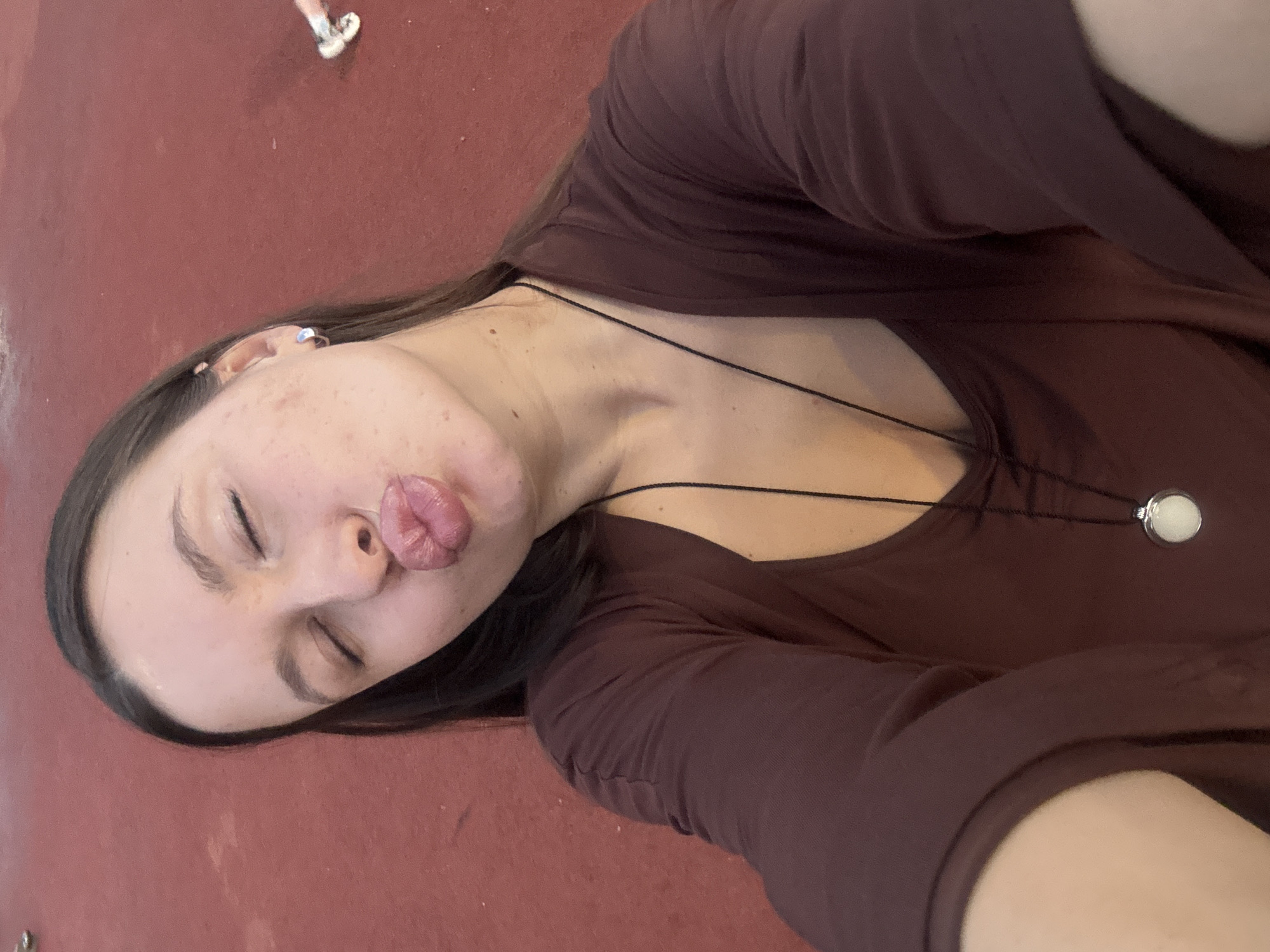 A woman lies on a red surface, possibly concrete or painted ground, with her eyes closed and lips pursed in a kissing gesture. She wears a dark maroon long-sleeved shirt and a delicate necklace with a circular pendant. Some hair is visible, dark and long. Another person's feet, shod in white shoes, are partially visible in the top left corner of the image. The location cannot be determined from the image alone, and there is no explicit information to identify it as being in Vniissok, Russia.