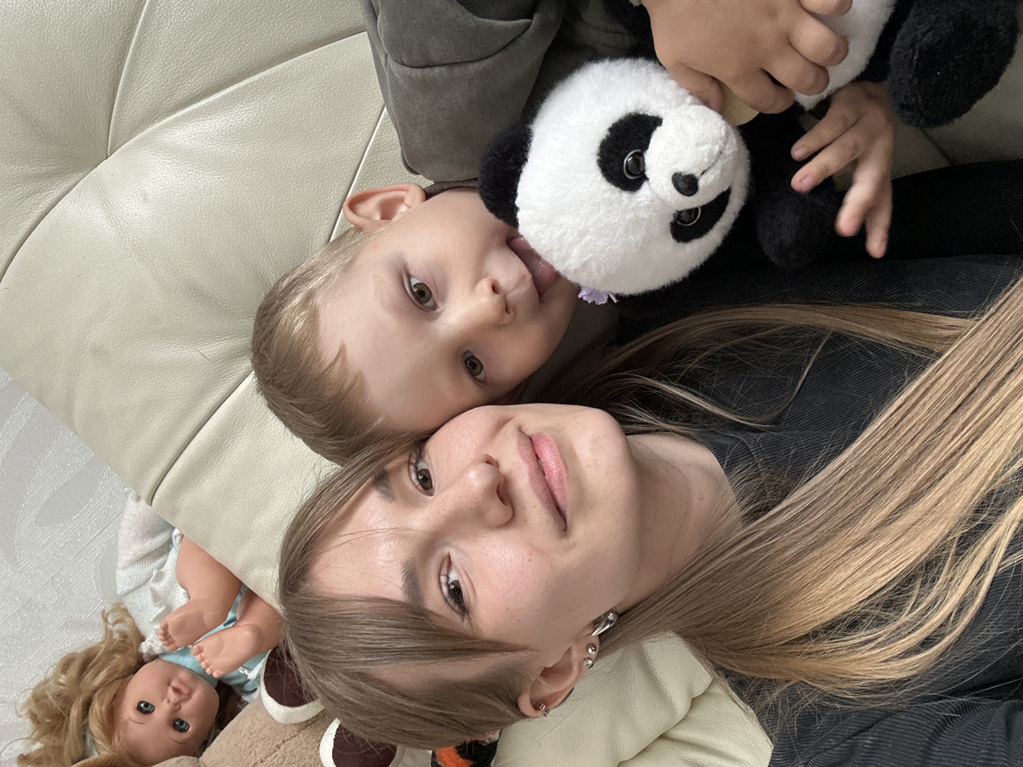 The image depicts a woman and a young child lying on a light-colored leather or faux-leather couch. The woman has light brown hair, fair skin, and is wearing earrings. The child, also fair-skinned, is holding a stuffed panda bear, and is sticking their tongue out. To the lower left of the frame, a doll with blonde hair and blue eyes is visible. The woman and child are close to each other and facing the camera. The location in the city of Sysert, Russia is not identifiable.