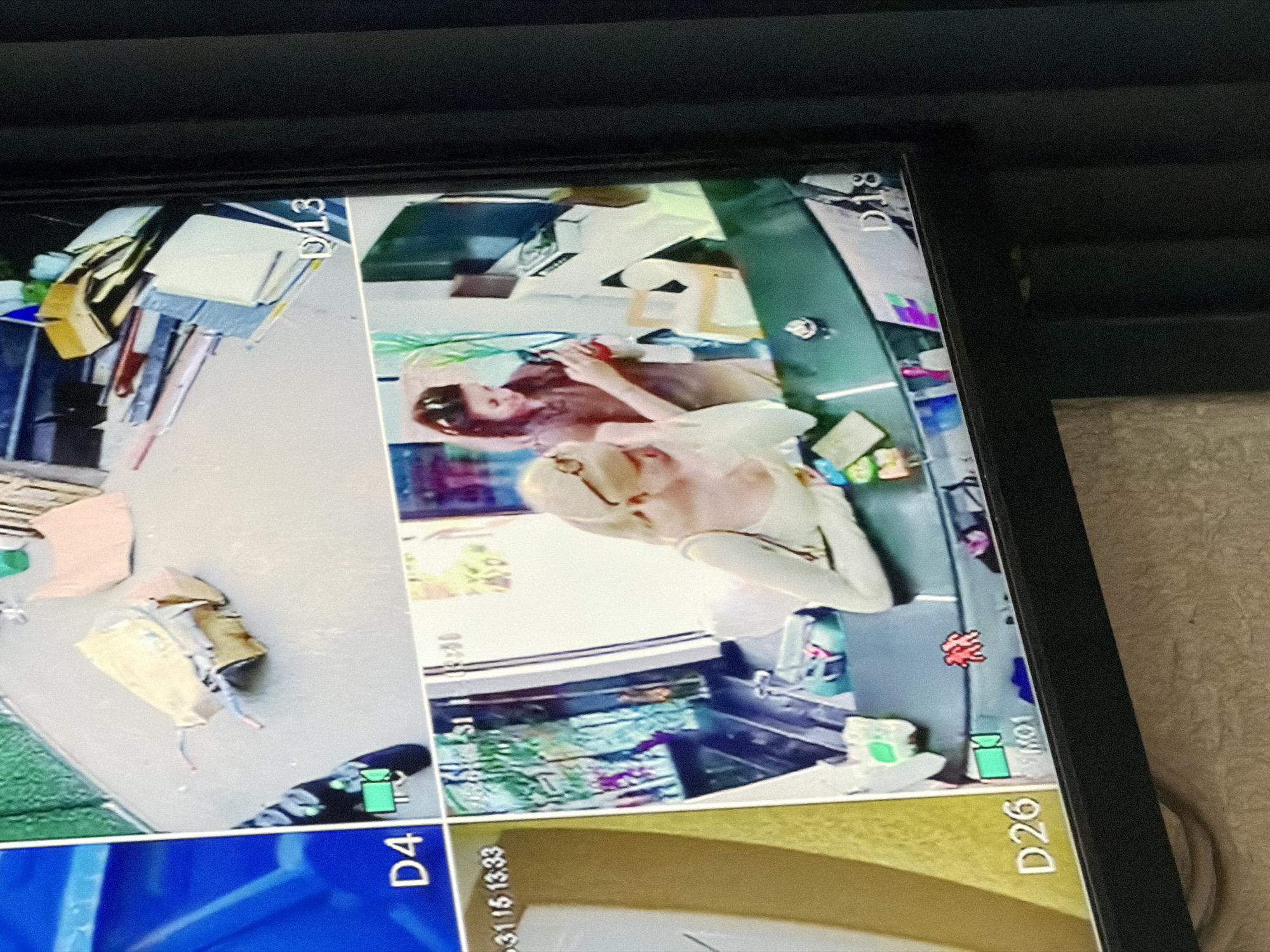 The image shows a surveillance monitor displaying four camera feeds. One feed shows a person lying on a table with another person standing next to the table. Another feed shows a blue bin. Another shows boxes and clothing items on a concrete surface. The image also shows text on the monitor: D13, D18, D4, D26 and numbers ending in 13:33