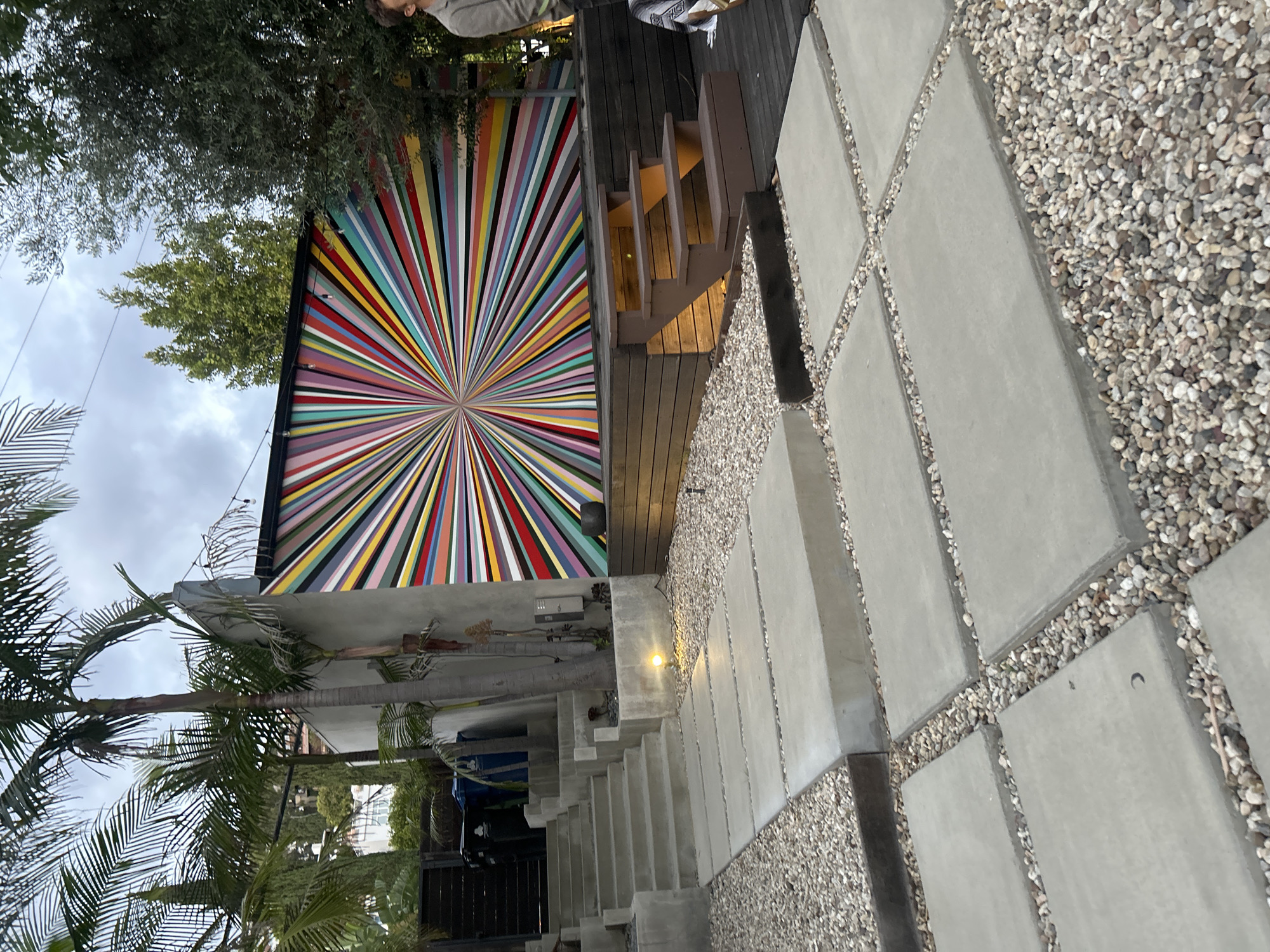 This image depicts a building exterior, featuring a large, colorful mural with a radial design of painted stripes emanating from a central point. The mural is installed on a light-colored concrete wall. Adjacent to the mural, there's a dark wooden structure with stairs leading up to a deck. The foreground consists of a pathway made of rectangular concrete slabs separated by gravel. The building appears to be in a residential area. The presence of palm trees suggests a warm climate, consistent with Southern California. Based on the architectural style, vegetation, and location, it is reasonable to infer that the image was taken in Los Angeles, California.
