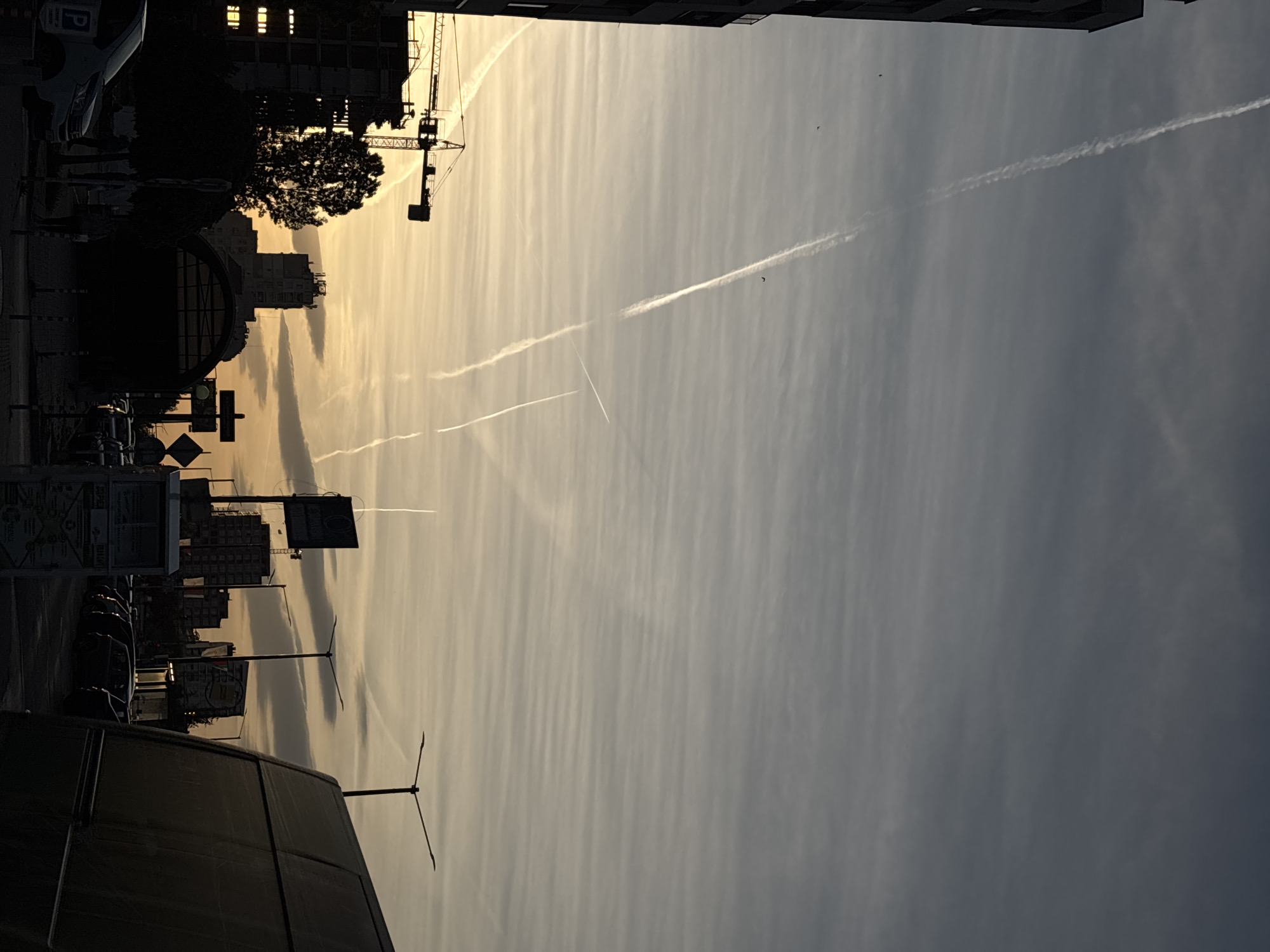 The image depicts a view of Sofia, Bulgaria, at dusk or dawn. The sky is striated with thin clouds and several contrails from airplanes. The silhouettes of buildings, trees, and a crane are visible against the lighter sky. A street scene is also present, including a car, street signs, and lamp posts.