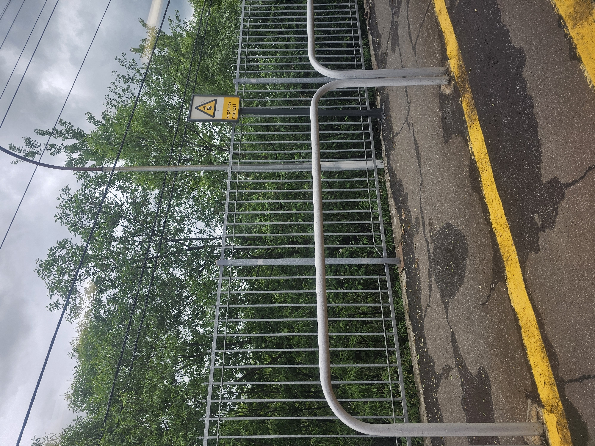 The image presents a scene near a railway line in Krasny Bor, Russia. A grey metal fence runs parallel to the tracks, likely as a safety barrier. A yellow line is painted on the asphalt ground marking the edge of the pathway or platform. Behind the fence, lush green trees and foliage are visible. An overhead power line stretches across the top of the frame. Affixed to the fence is a yellow warning sign, displaying a black silhouette of an electrical hazard and text in Cyrillic. The sky appears overcast and cloudy. The asphalt surface shows some signs of wear or stains. There is no presence of people or animals.