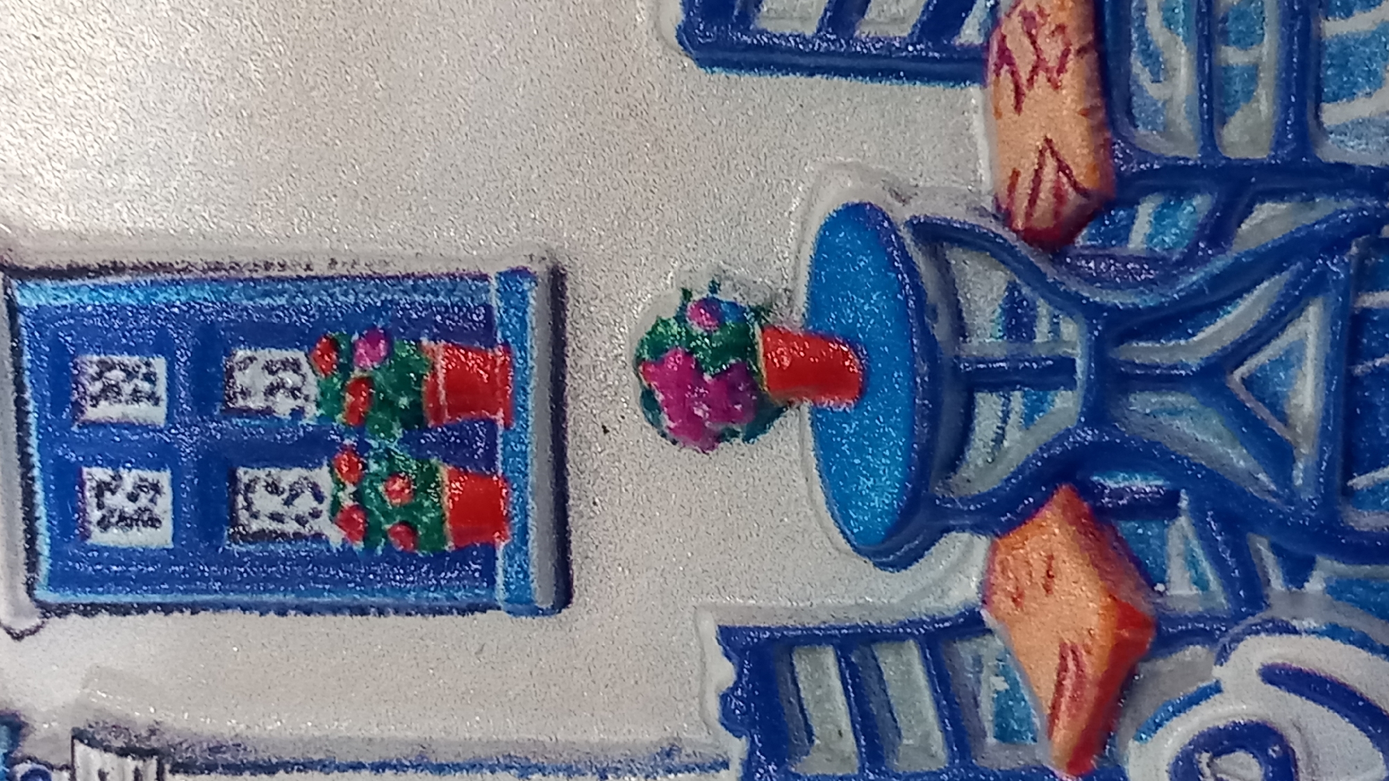 This is a close-up, overhead, low-angle shot of a miniature, glossy, three-dimensional representation of a typical scene from the city of Kos, Greece. The scene depicts a small, blue table with a red vase holding a pink and green floral arrangement. Two blue chairs, one on either side of the table, have orange cushions. In the background is a blue window with white panes and two red flower pots, each with green plants and red flowers. The overall aesthetic is reminiscent of a souvenir or decorative item.