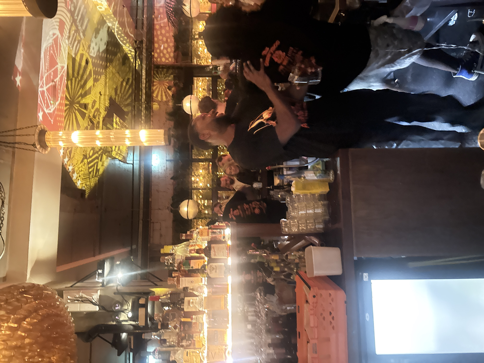 Here is a description of the image:
**Scene:** Interior of a bar, likely in London, United Kingdom. The scene is dimly lit.
**People:** Several people are present, including two individuals positioned in the foreground, engaged in an interaction. It is hard to see their facial expressions. Other people, likely bar patrons, are in the background. The person in the foreground is dressed in a dark T-shirt with a graphic on it.
**Objects:** A long bar stretches across the scene, with numerous bottles of alcohol lining the back. Glasses, drinkware, and other bar equipment are visible on the bar surface. There are many lights, and decorative light fixtures hanging from the ceiling. There is a large blank white rectangle.
**Actions:** The two people in the foreground appear to be engaged in a close interaction. One is leaning in towards the other, who has their hand on the back of the first person, suggesting they are holding them. The lighting in the scene is very soft and inviting.
**Environment:** The bar has a casual and possibly trendy vibe. The lighting and decor are designed to create an inviting and social atmosphere.