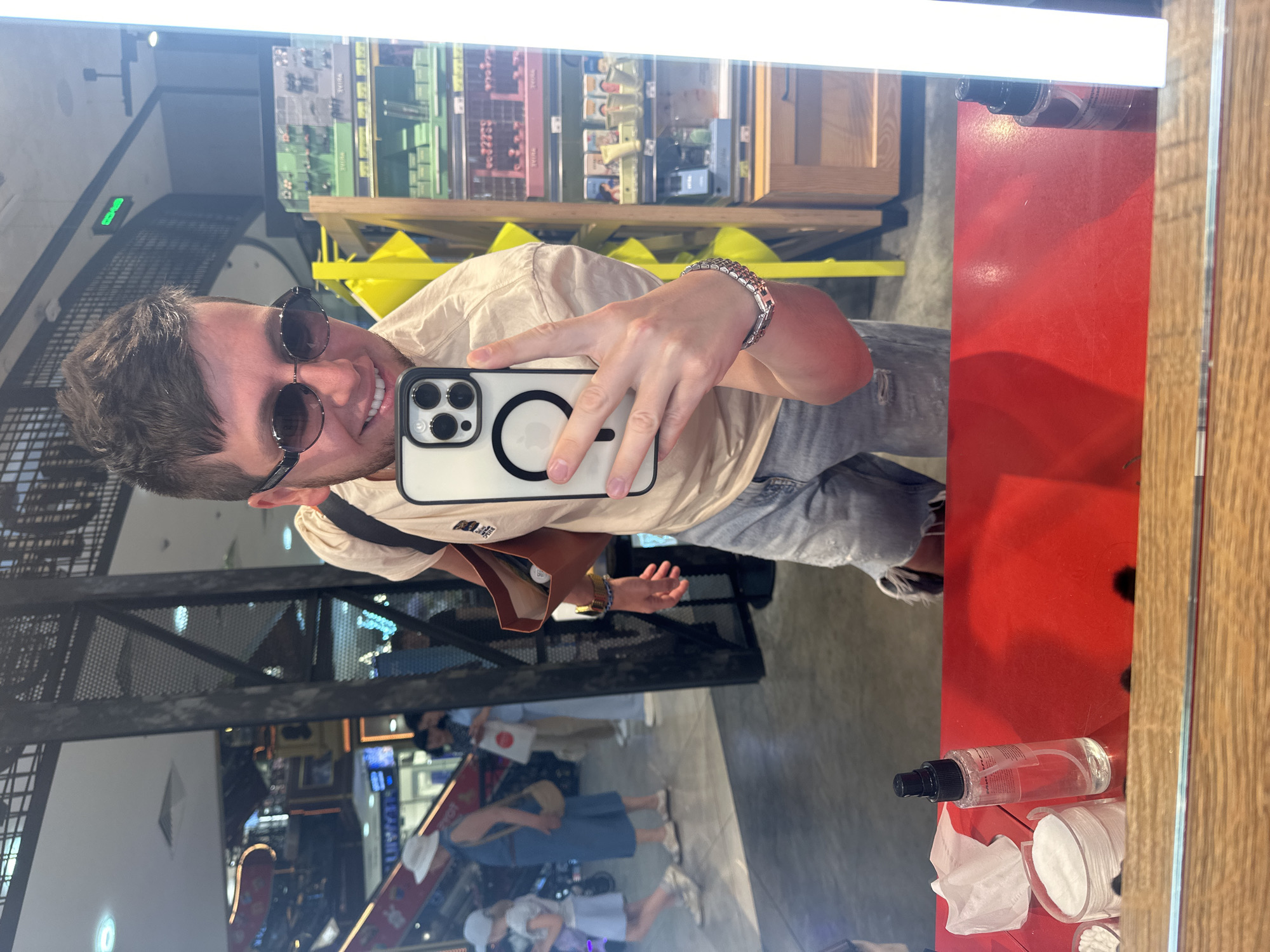 A man takes a selfie in a mirror, wearing sunglasses, a beige t-shirt, and ripped denim shorts. He has a brown bag over his shoulder. In the background, the mirror reflects a store interior with shelves stocked with products, escalators, and other shoppers. The scene appears to be a retail environment within a shopping mall.
