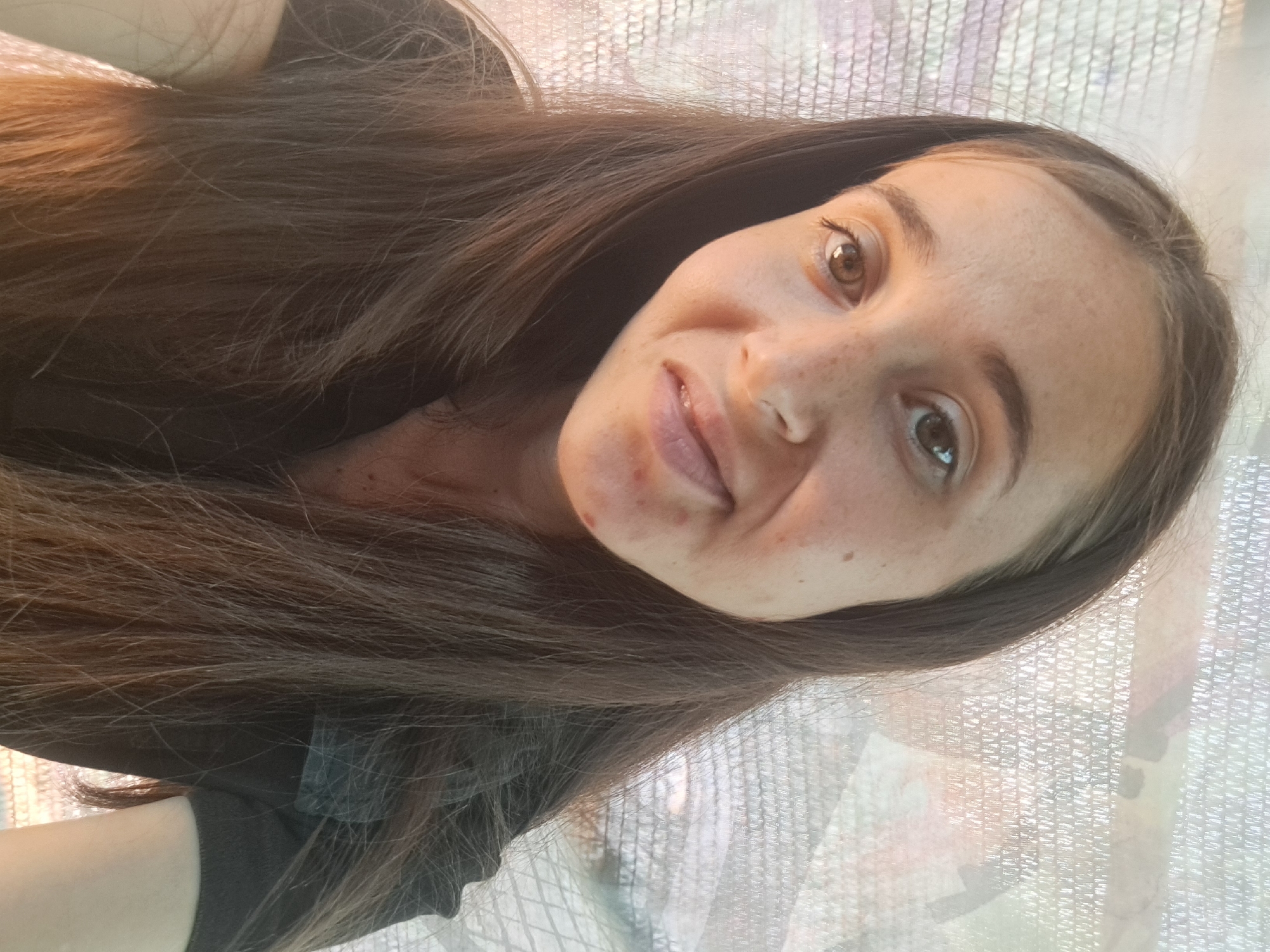 A woman with long brown hair and light skin is shown in a close-up, head-and-shoulders portrait. She has brown eyes and a smattering of freckles across her face. Her lips are slightly parted, and she has a subtle smile. She is wearing a dark-colored shirt. The background appears to be a netted or screened surface, possibly a shade cloth, with hints of abstract color and light filtering through. The image was taken in Vypasne, Ukraine.