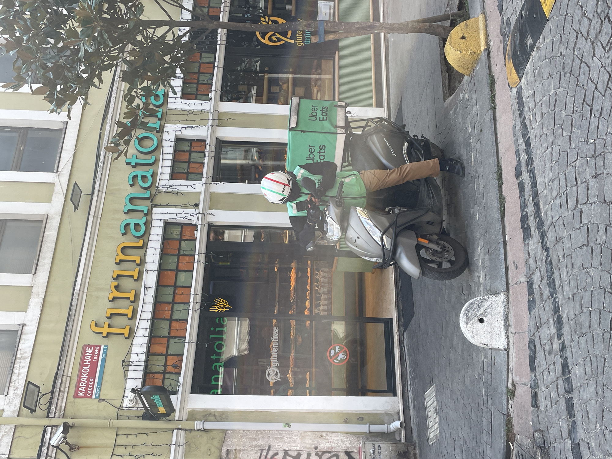 A delivery person wearing a helmet with Italian flag colors and a green vest emblazoned with "Uber Eats" is seated on a scooter outside a bakery in Kadıköy, Turkey. The scooter has a large green Uber Eats delivery box on the back. The bakery, named "Fırın Anatolia" and featuring a "gluten free" sign, is adorned with string lights and decorative windows. The street is paved with cobblestones, and a white bollard stands on the sidewalk. The scene appears to be a typical urban streetscape in Turkey, with a focus on the modern delivery service operating in a traditional setting.