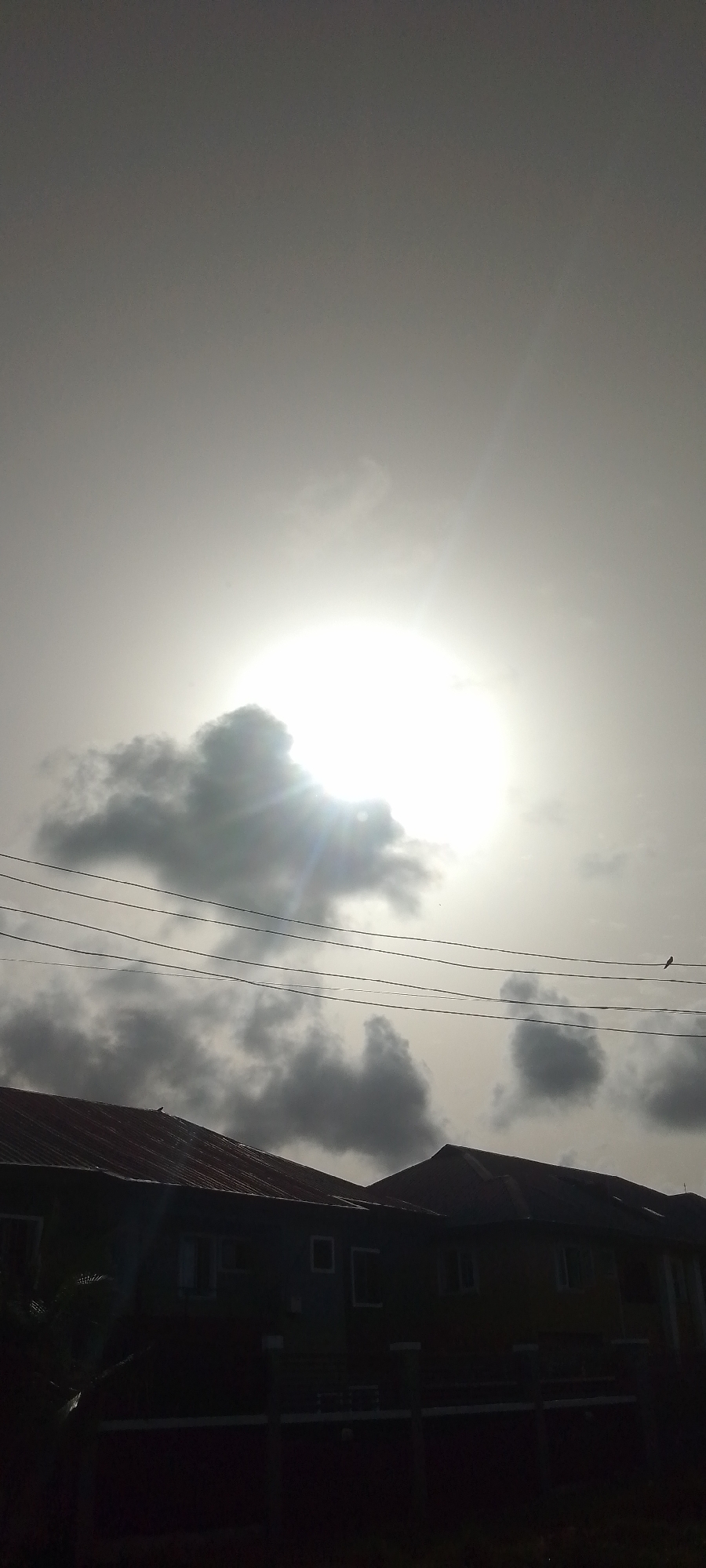 The image depicts a sky with the sun obscured by clouds, emitting light rays. Below, a row of buildings with metal roofs is visible, and electrical wires stretch across the scene. The location is Unknown, Unknown.