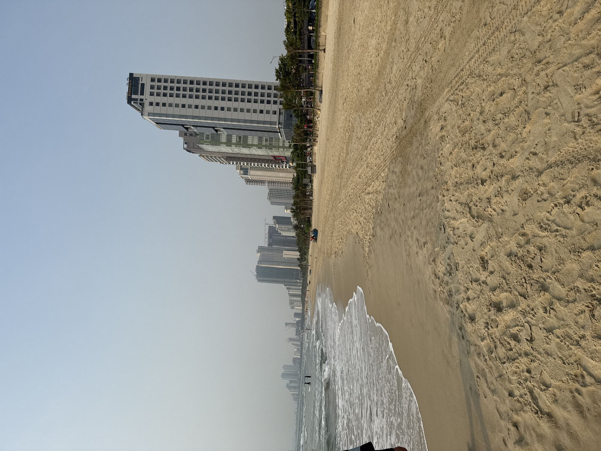 The image shows a beach scene in Đà Nẵng, Vietnam, with the cityscape visible in the background. The beach features light brown sand with wave patterns and tire tracks. The ocean is light blue with visible waves approaching the shore. Several individuals can be seen in the water. Along the shoreline, a line of palm trees and buildings forms the city's edge. Skyscrapers dominate the urban landscape, with a prominent rectangular building labeled "Peninsula" on its top edge. The sky is a clear, pale blue.