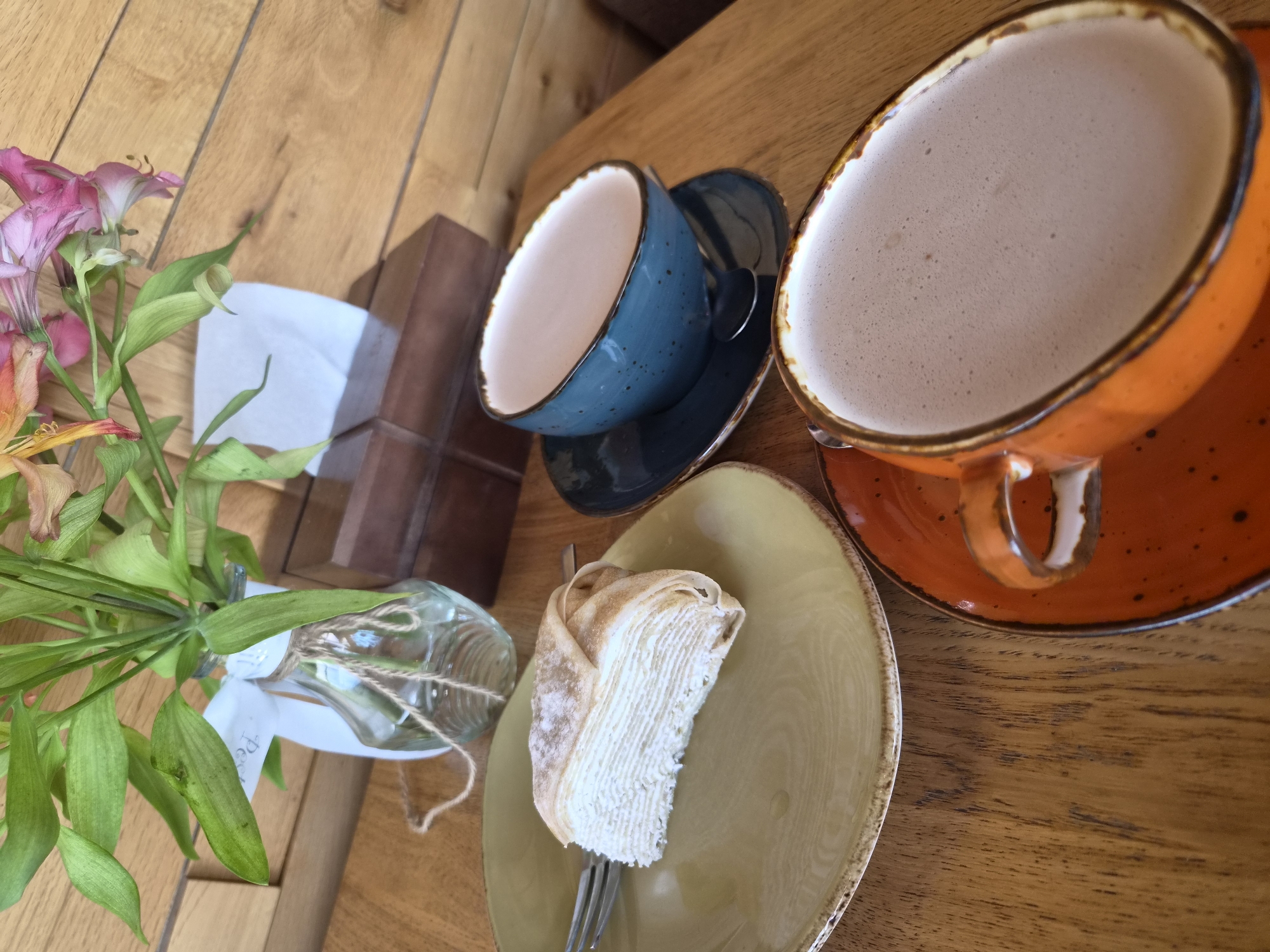 The image depicts a close-up, slightly overhead view of a wooden table with a dessert and two cups of coffee. On a light green plate, a slice of layered crepe cake is dusted with powdered sugar, with a fork resting to the left. To the right of the cake, a blue ceramic cup sits on a dark blue saucer, containing a frothy beverage. Further to the right and closer to the foreground, an orange ceramic cup on a matching saucer holds a similar frothy beverage. In the background, a bouquet of flowers in a clear glass vase with twine and a white ribbon is visible, along with a dark wooden tissue box. The overall setting suggests a cafe or restaurant environment. This image was taken in Arseniev, Russia.