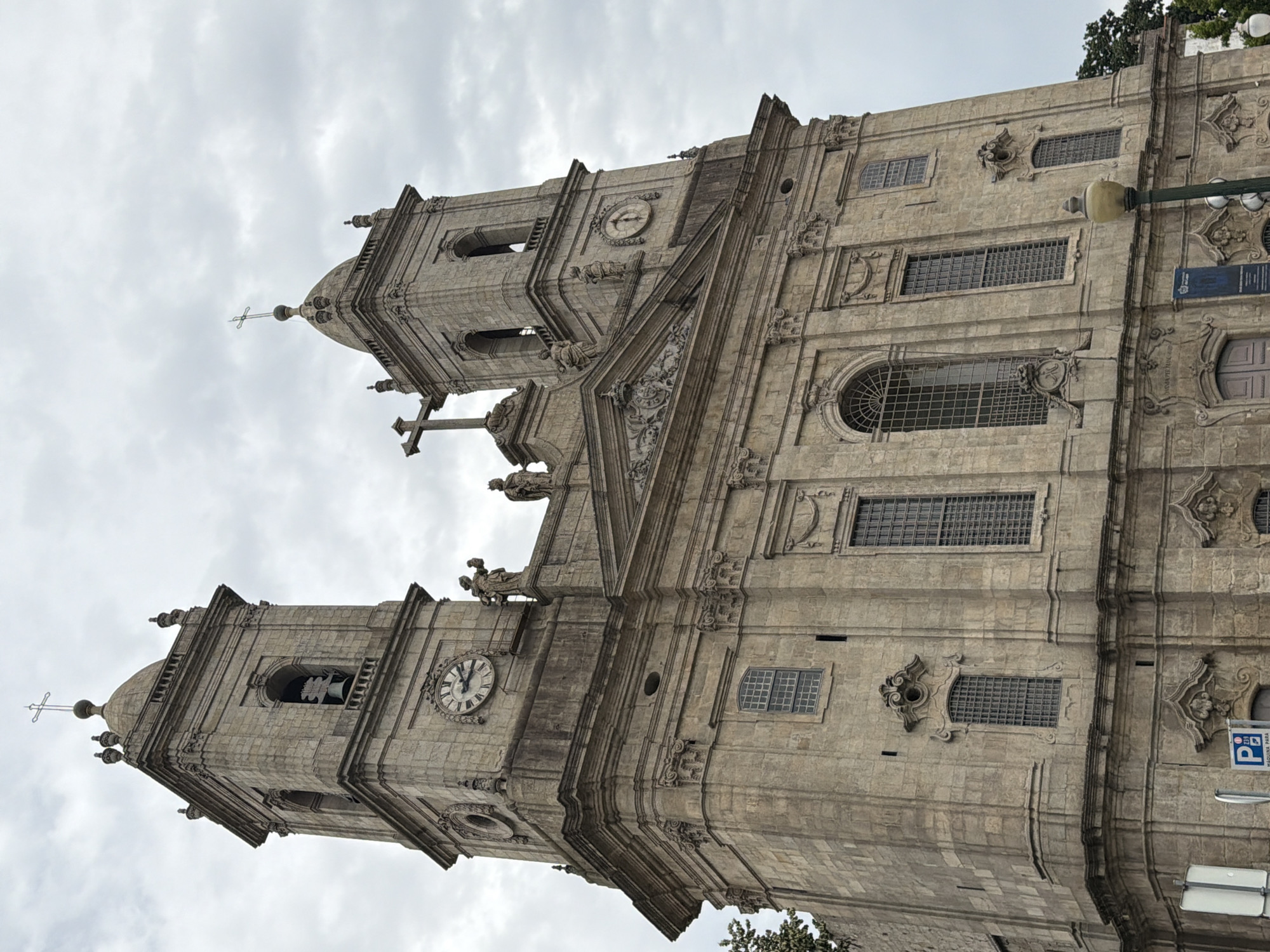 This is a full-shot, eye-level, exterior, daytime photograph of the Igreja dos Clérigos (Clérigos Church) in Porto, Portugal. The sky is overcast with a medium-grey cloud cover. The church is constructed of stone and features two tall bell towers, each topped with a dome and a cross. A large clock face is visible on the left tower, and a smaller clock face is on the right tower. There are decorative sculptures of figures on the facade, and numerous windows. The lower part of the church shows a large entrance door with arched windows above it. A street lamp is visible in the lower right corner of the image, and a partial view of a street sign is in the lower left corner. There are no people visible in the image.