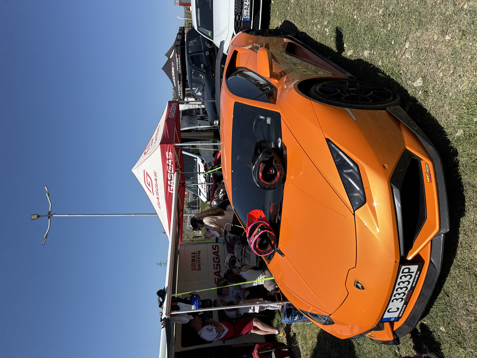 The image shows an orange Lamborghini parked on a grassy surface under a clear blue sky. A helmet rests on the car's windshield. In the background are tents and vehicles. The tents have "GASGAS" printed on them. One of the vehicles has "AIRMAX БЛАГОЕВГРАД" printed on the side. A license plate "C 33333P" is visible on the front of the Lamborghini. Other people are visible near the tents and vehicles. The overall scene suggests an outdoor event or gathering in Selishte, Bulgaria.