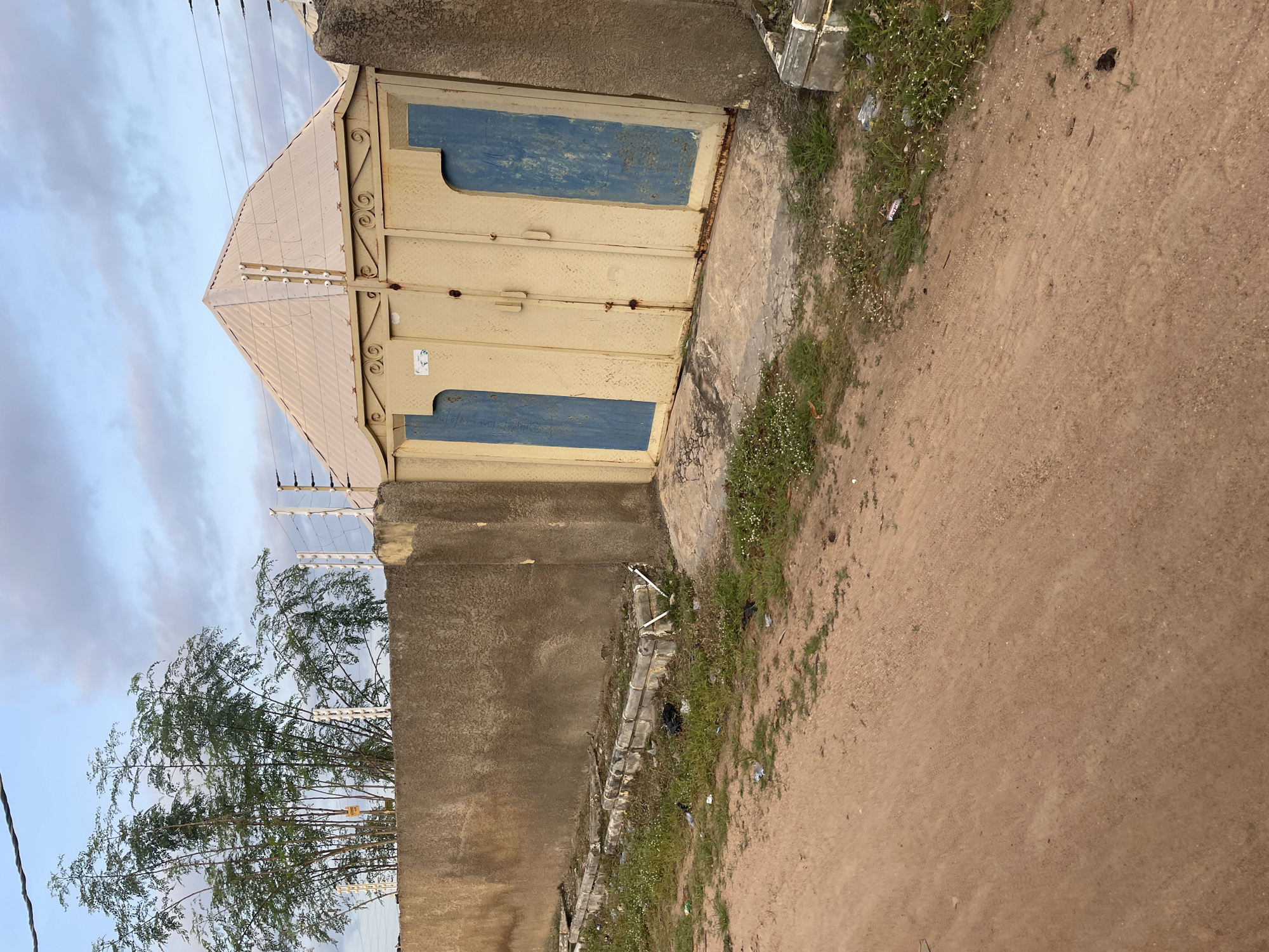 The image depicts a scene on a dirt road in Jalingo, Nigeria. In the foreground, the road is made of reddish-brown soil, with sparse green grass and some white flowering weeds growing along the edges. A concrete curb separates the road from a patch of overgrown grass littered with debris, including plastic bags and papers.
To the right and extending into the background is a tall, rough-textured concrete wall, painted a yellowish-brown hue on its right side. Adjacent to this wall is a large, imposing metal gate, painted yellow with two vertical blue rectangular panels on each leaf. The gate has a decorative, wavy metal trim along its top edge. Above the gate, a sloped, corrugated metal roof of a building is visible, with electric fence poles mounted along its peak.
To the left of the gate and extending behind the wall, a cluster of tall, slender trees with green foliage is present. The sky above is partly cloudy with a mix of light blue and grey clouds. Power lines are visible spanning across the sky. The overall impression is one of an exterior urban or semi-urban environment in Jalingo, Nigeria.