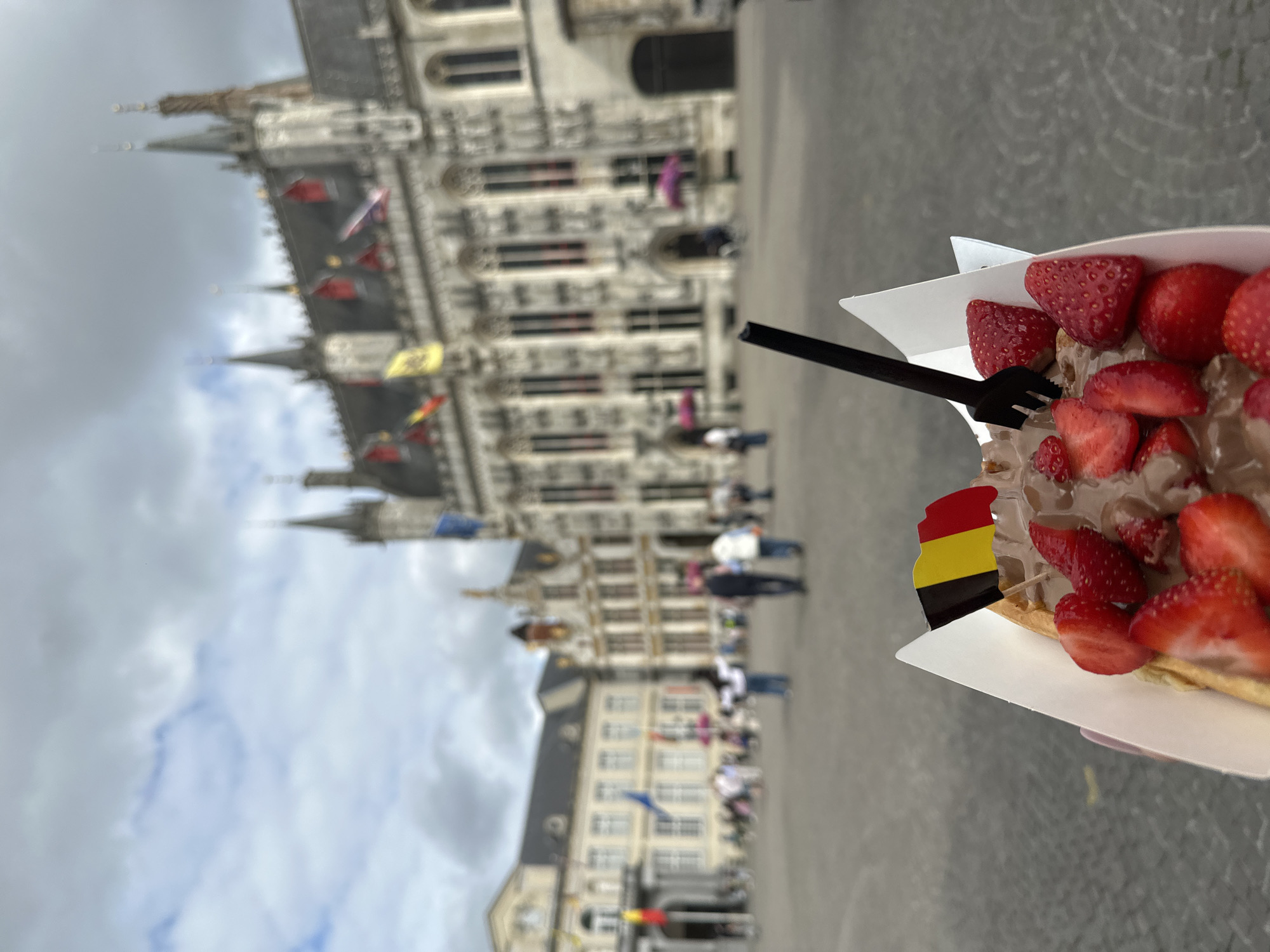 An outdoor, eye-level, medium shot shows a Belgian waffle topped with chocolate sauce and sliced strawberries, held in the foreground. A small Belgian flag is stuck into the waffle. A black plastic fork is inserted into the waffle, and the paper container holding the waffle is partially visible. In the background, the blurry outline of the Bruges city hall, the Belfry of Bruges, and other historic buildings with Gothic architecture is visible. The sky is cloudy. People are walking in the square in front of the buildings.