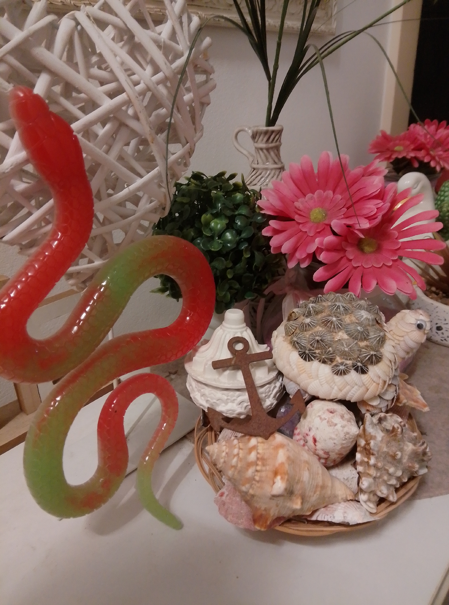 The image depicts a decorative arrangement on a white surface, likely a table. In the foreground, a long, red and green, segmented, serpent-like object snakes across the scene. To the right of the serpent, a woven basket holds a collection of seashells and a decorative turtle made of shells. Behind the basket and to the left, a white, ornate object with a small anchor resting on it is visible. Further in the background, pink artificial flowers are arranged with greenery. A large, white, woven heart-shaped object is positioned on the left side of the image. The scene is brightly lit, suggesting an indoor setting, possibly a home or a shop display. There is no indication of the city of Wedel, Germany in the image.