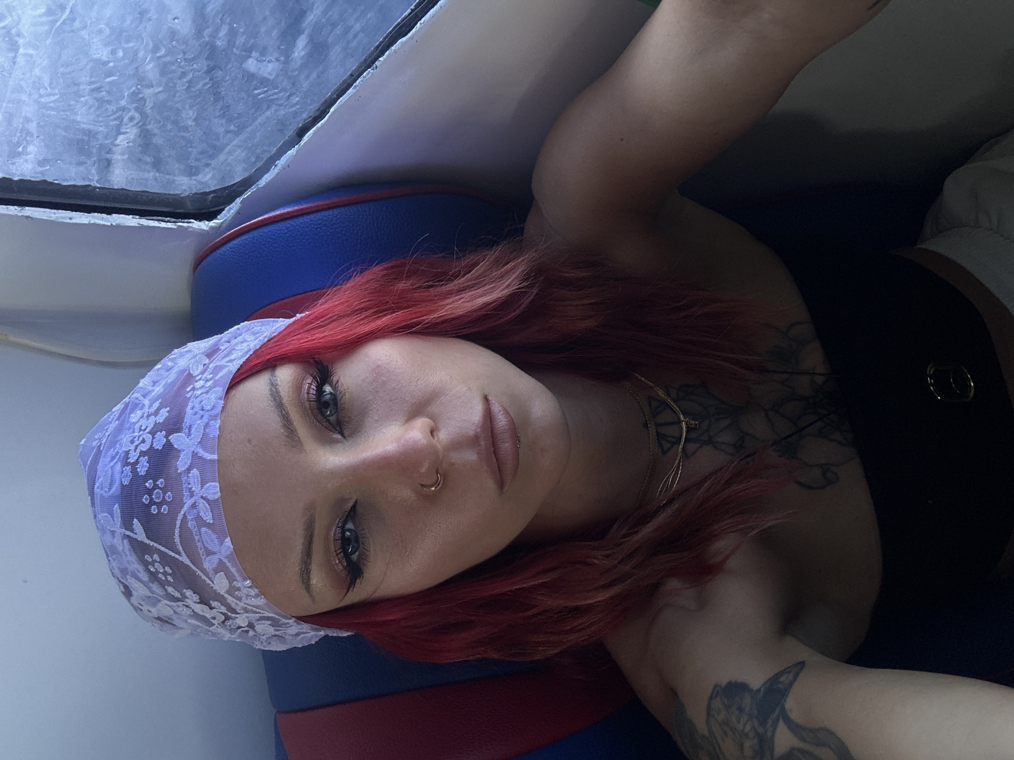 A woman with red hair and a white lace headscarf poses for a selfie indoors. She has tattoos on her chest and arm. The background appears to be the interior of a vehicle or boat with blue and red seating. Location is not discernible based on the image.