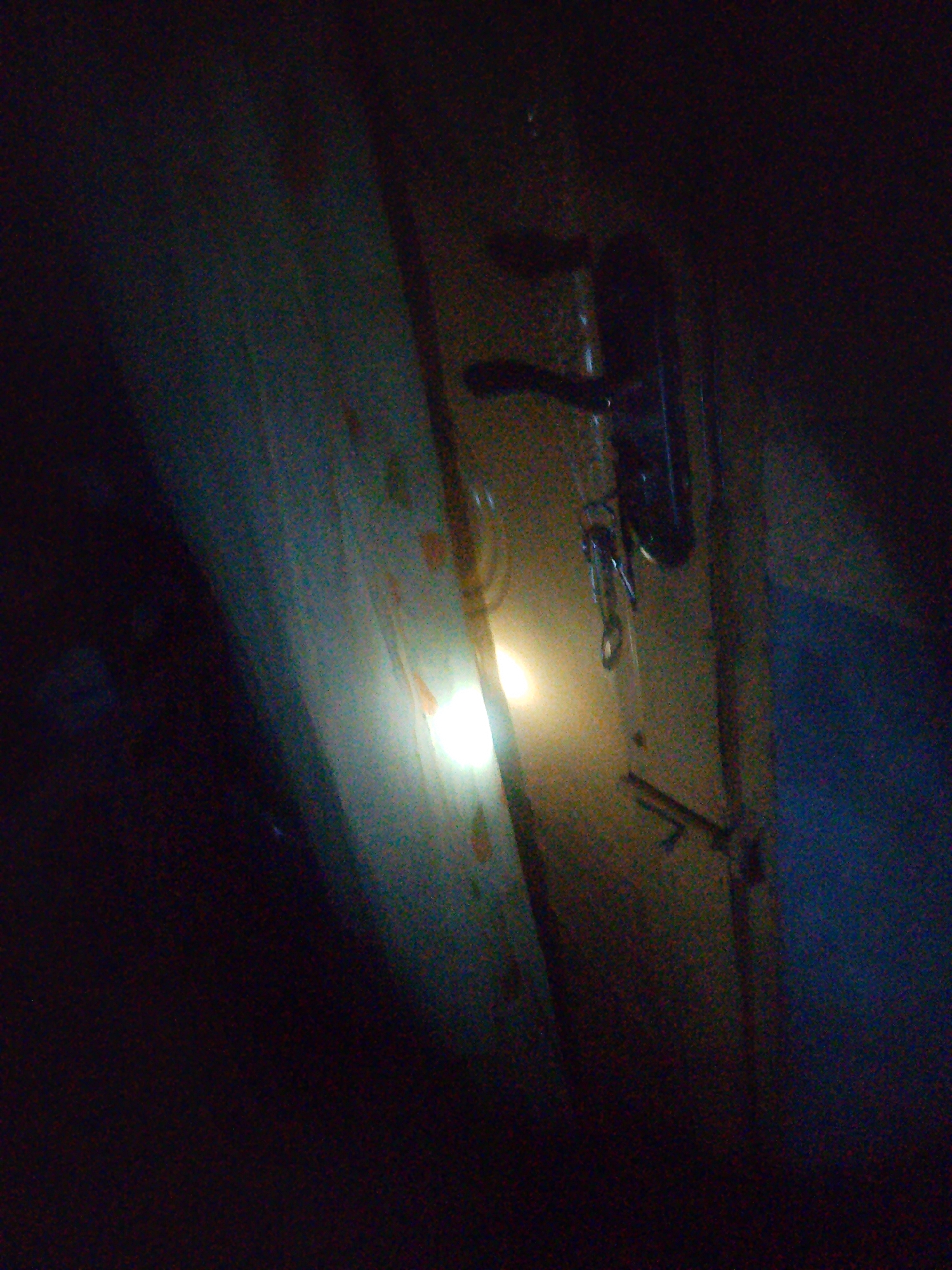 The image depicts a dimly lit scene, likely indoors. The primary subject is a door handle, which appears to be made of metal and is attached to a wooden door. A keychain with a visible ring is attached to the handle. To the left of the door, a curtain or fabric is visible, partially illuminated by a bright light source that is out of frame. The overall impression is one of low light conditions, possibly at night or in a poorly lit room. There is no discernible text within the image. The location context of Jalingo, Nigeria, does not provide specific visual cues within this image, as it is a generic interior shot.