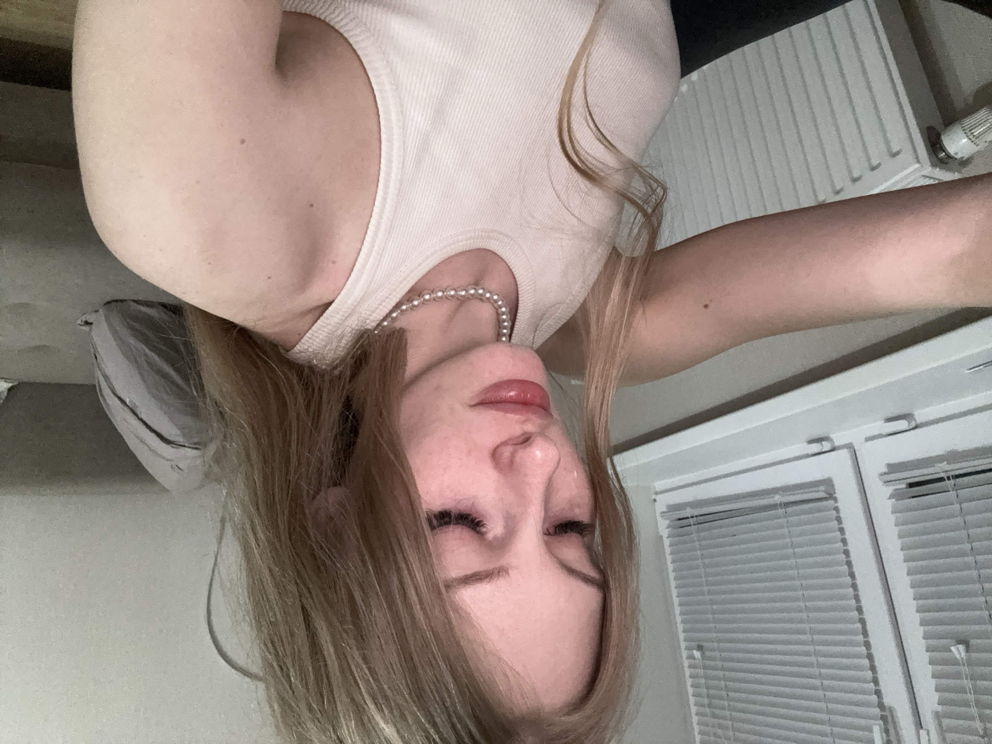 The image shows a young Caucasian woman with blonde hair and fair skin. She is wearing a white tank top and a pearl necklace. Her eyes are closed and she appears to be crying. The background shows a window with blinds and a radiator, suggesting an indoor setting. The overall mood of the image is somber and intimate. The image does not contain any information that would indicate the location within the city of Perm, Russia.
