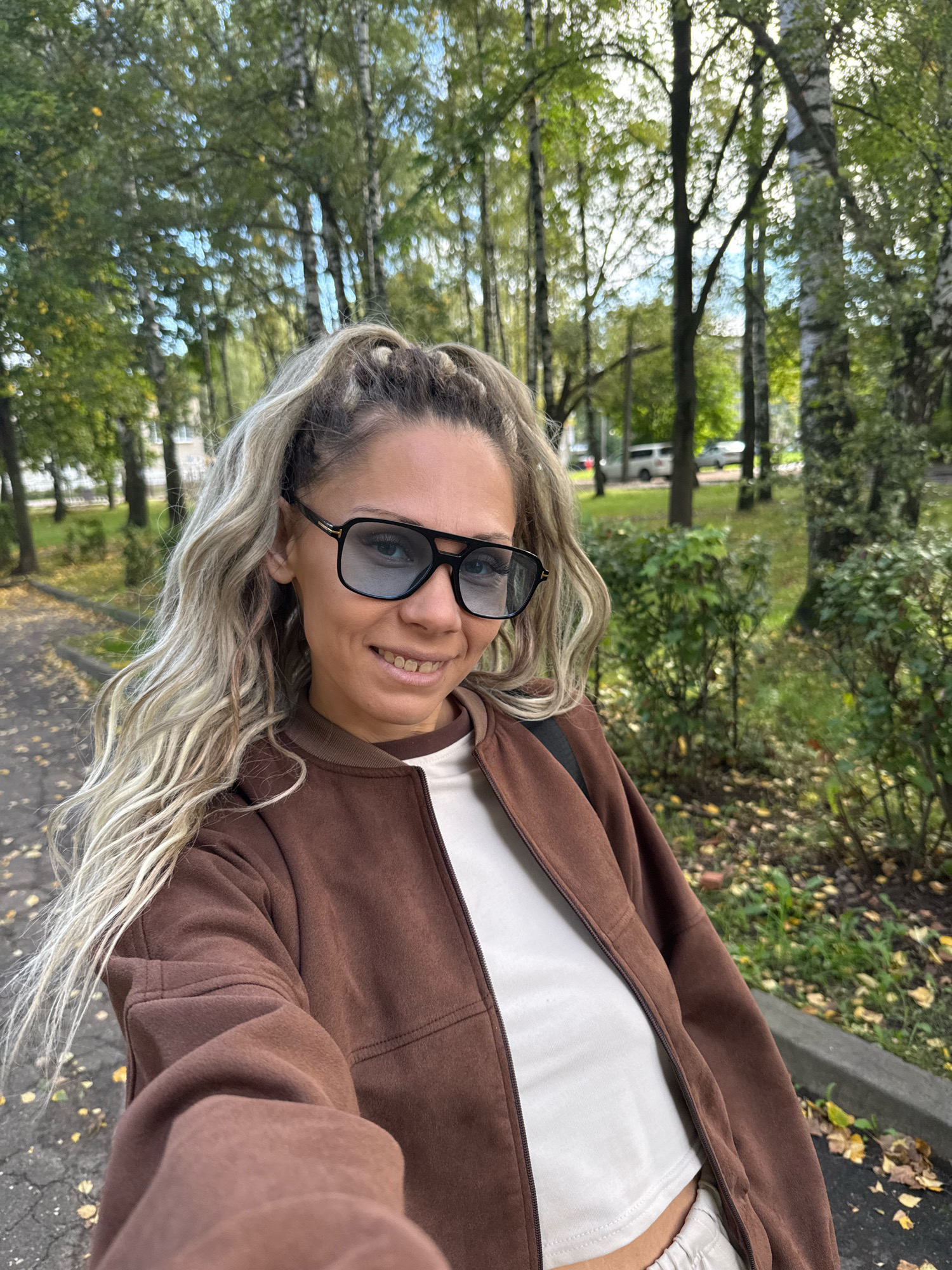 A woman with blonde hair pulled into a high ponytail and wearing sunglasses and a brown jacket stands in an outdoor urban park. She is smiling and looking at the camera. The background shows trees, green bushes, and a paved walkway with fallen leaves. In the distance, a few cars are parked along a street.