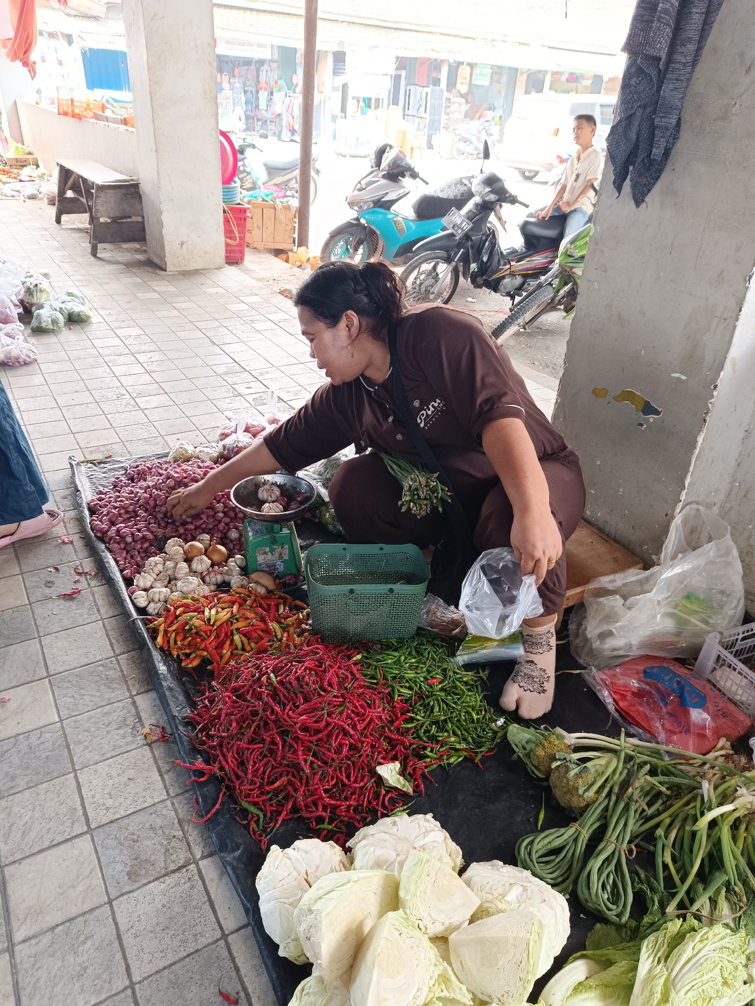 A woman is selling vegetables in a market in Kayu Agung, Indonesia. She is kneeling on the ground, surrounded by a large assortment of produce including red onions, garlic, chilies, green beans, and cabbage. A green weighing scale is visible near the onions. In the background, there are motorcycles and other market stalls. The woman is wearing a brown shirt and has her hair tied back. She is holding a plastic bag and appears to be in the process of weighing or packaging some items.