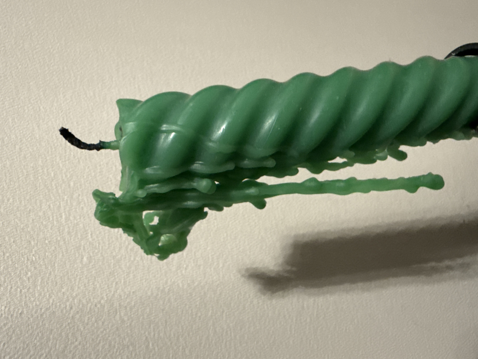 The image displays a green, twisted candle, positioned horizontally against a light-colored, textured wall. The candle shows clear signs of having been partially burned, with a short, black, charred wick visible at its left end. Substantial amounts of melted green wax have dripped from the candle's body, solidifying into irregular, organic shapes and strands beneath it, adhering to the main candle structure. Some of these wax drippings extend horizontally, creating elongated forms. The twisted pattern of the candle is prominent along its length.
A soft, elongated shadow is cast on the wall below the candle and its wax drips, indicating an external light source. The background wall is uniform in color, likely an off-white or light beige, and exhibits a subtle, coarse texture. No other objects, people, or discernible text are present in the frame. The image focuses solely on the candle and its immediate surroundings. The scene depicts the aftermath of a candle burning, showcasing the solidified wax flow. The provided location context for this media is Berezyna, Ukraine, although no specific geographical cues are visible within the image itself.