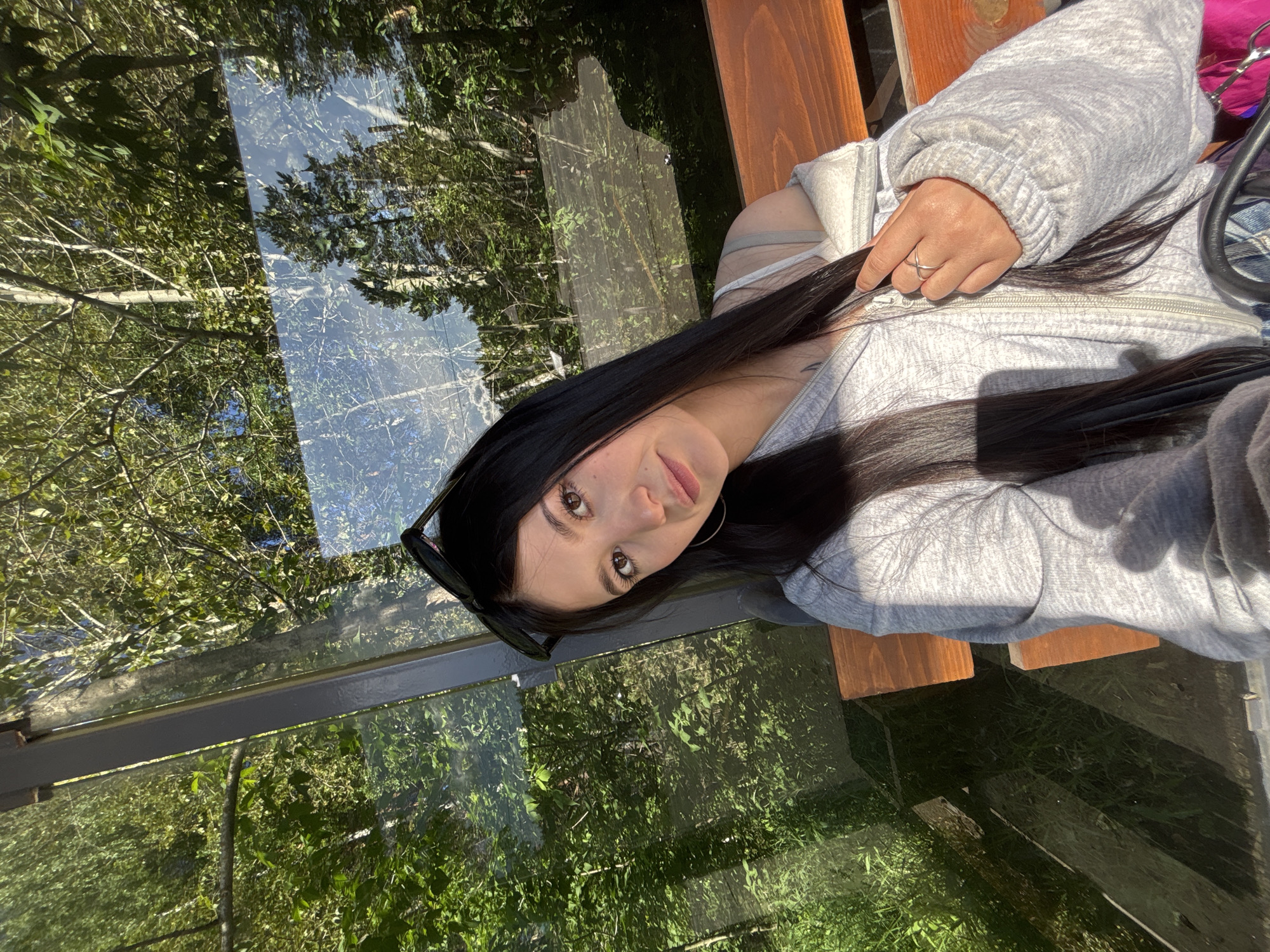 A young woman with long, dark hair and light brown eyes is taking a selfie outdoors. She is wearing a grey zip-up hoodie over a tank top, and sunglasses are perched on her head. A silver ring is visible on her right hand. She is sitting on a wooden bench next to a reflective glass structure, possibly a bus stop shelter, that mirrors the surrounding green trees and blue sky. The setting appears to be a park or natural area within the city of Chaikovsky, Russia, indicated by the presence of urban infrastructure alongside lush greenery. The lighting suggests it is daytime, likely late afternoon given the warm tones and shadows.