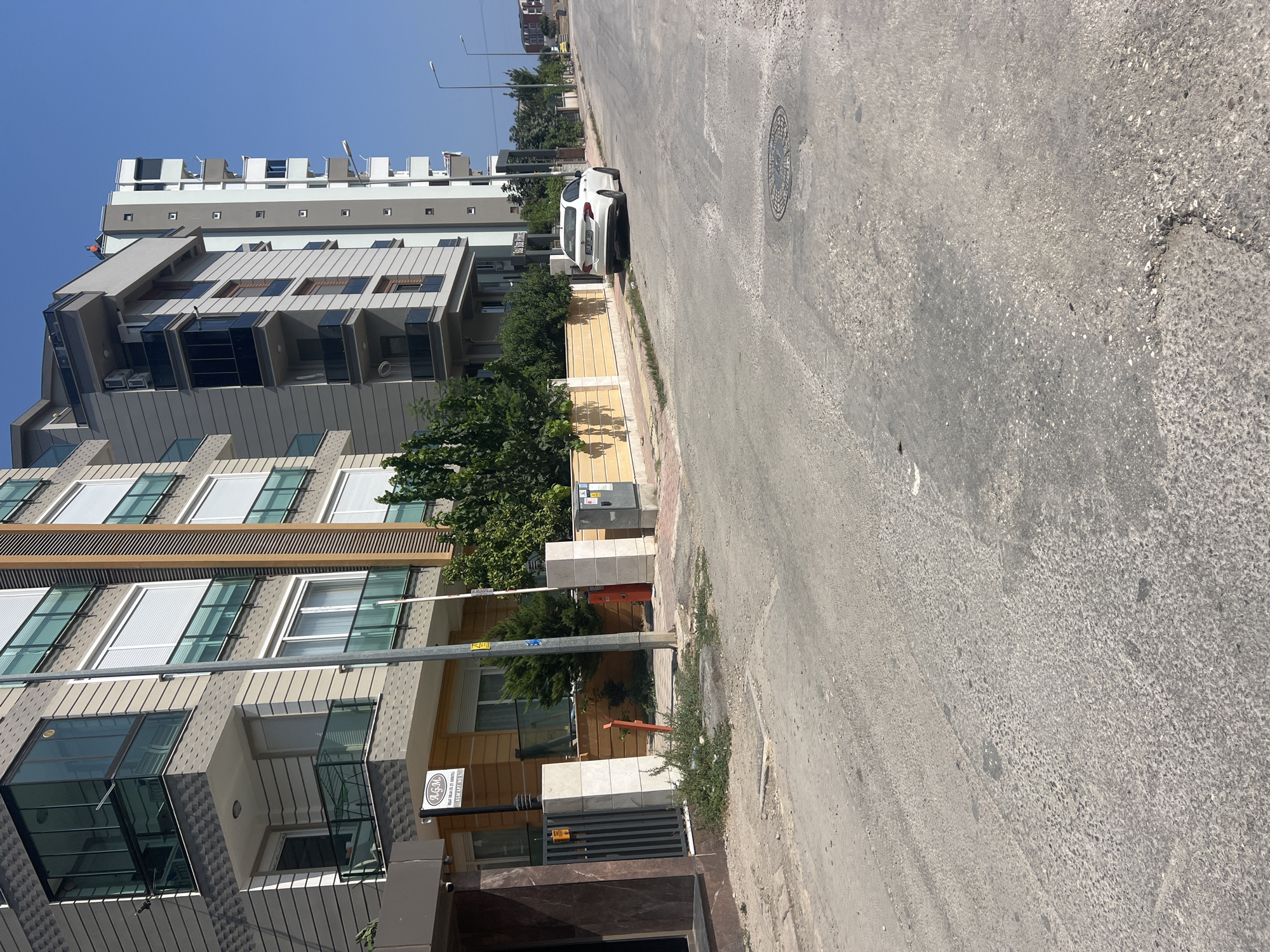 A white sedan is parked on the side of a paved street in Konyaaltı, Turkey. The street is lined with modern apartment buildings and trees. The sky is clear and blue.