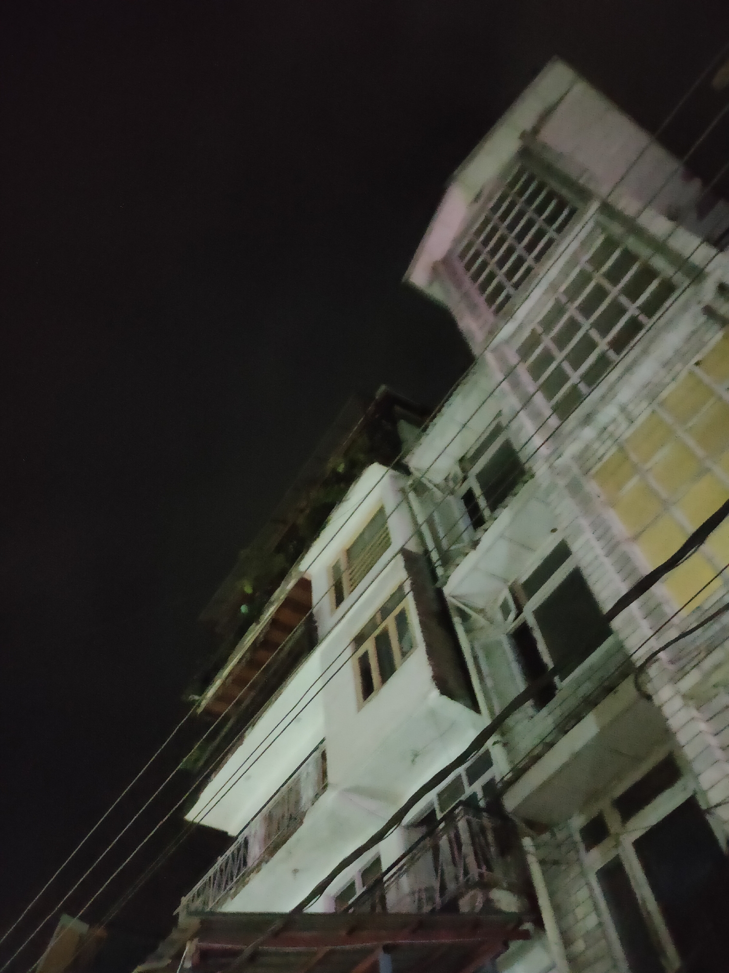 The image shows a multi-story building at night in Manali, India. The building appears to be made of concrete and has several floors with balconies and numerous windows. The windows are mostly rectangular, some with grid-like frames. The building's facade is painted white, with some sections showing yellowed or stained areas. Electrical wires are strung across the front of the building. There are no people visible in the image. The sky is dark, indicating nighttime.