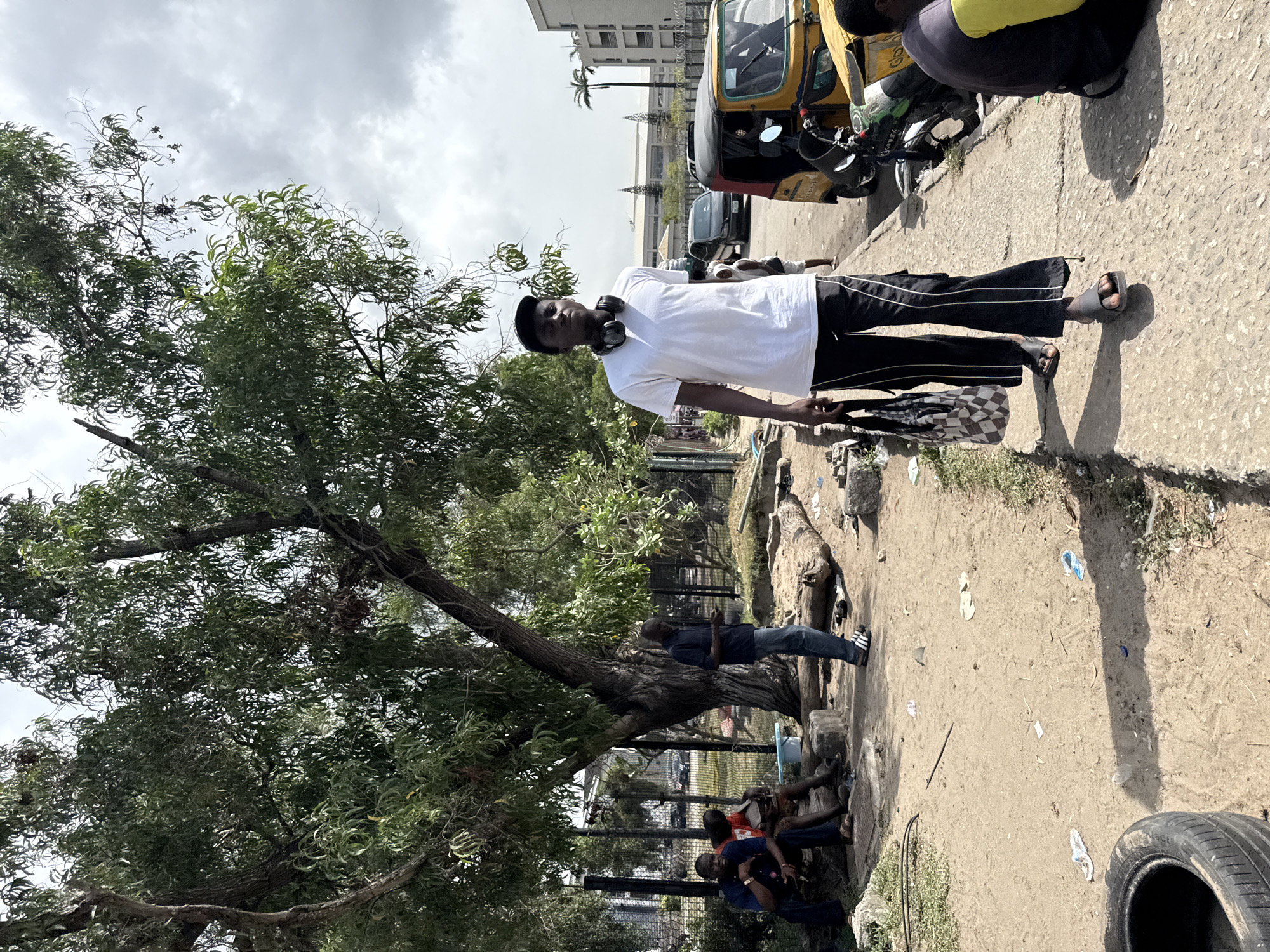 This image depicts an outdoor urban or semi-urban scene in Lekki, Nigeria, under an overcast sky with bright daylight suggesting midday or early afternoon.
In the foreground, a young man stands, looking directly towards the camera. He wears a white t-shirt, black track pants with white stripes, and black headphones around his neck. He carries a black and white checkered bag in his left hand.
To the left, a large tree with dense green foliage dominates the frame, casting shadows. Underneath and around the tree, several other individuals are present; one person in a blue shirt and jeans stands, while two others are seated, appearing to be resting or conversing.
On the right side of the image, a paved surface transitions into a dirt path. A yellow Keke (auto-rickshaw), a common mode of transport in Nigeria, is prominently parked in the midground, with a partially visible person sitting nearby. A motorcycle is also visible next to the Keke. In the background, further vehicles are discernable, along with distant buildings and a metal fence. The ground is scattered with various pieces of litter, including plastic bottles and papers. An old car tire rests in the bottom right corner.
The scene captures everyday life and interactions in a public space. No discernible text is visible in the image.