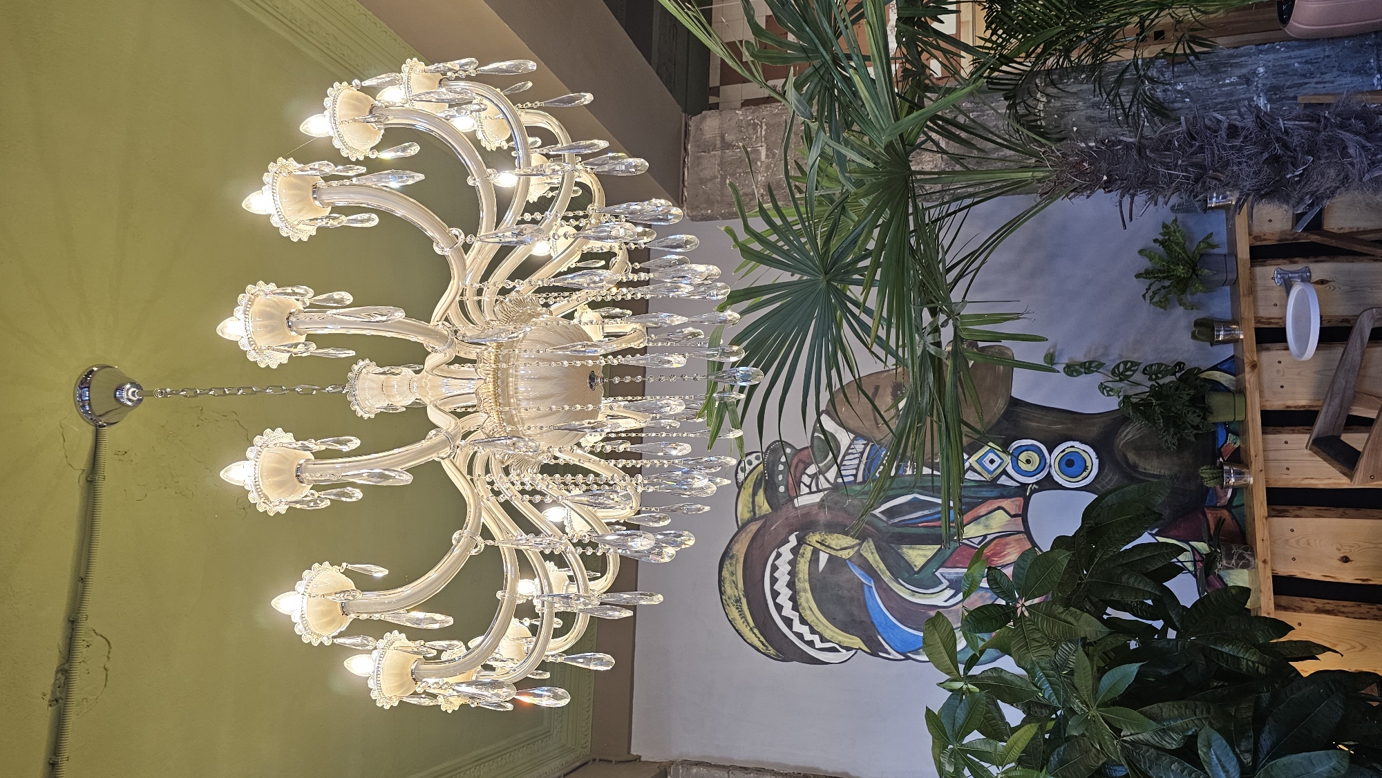 A close-up, low-angle interior shot captures a multi-tiered, ornate chandelier suspended from a pale green, textured ceiling. The chandelier is primarily white and adorned with numerous clear, faceted crystal drop pendants that catch and reflect light. Its structure consists of curved arms supporting exposed, bulb-shaped lights. Below the chandelier, a large, stylized mural depicts the profile of a person with dark skin, wearing a colorful, patterned headdress and large circular earrings. In the foreground, various green potted plants, including a palm tree with feathery fronds, are visible. The plants are arranged on a rustic wooden structure, which forms a shelf or counter. The background includes hints of concrete pillars and textured walls, suggesting an indoor, possibly commercial, space. The lighting is warm and ambient, emanating from the chandelier.