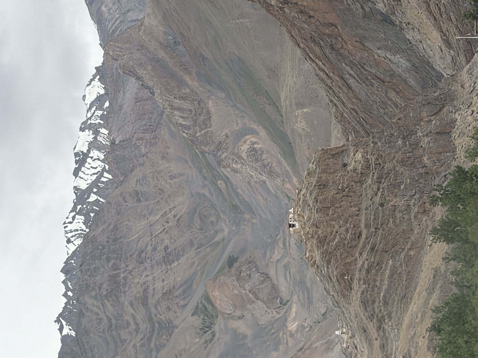 The image shows a wide, mountainous landscape under a cloudy sky. The mountains are rugged and appear to be barren with patches of snow on the higher peaks. In the mid-ground, perched on a rocky outcrop, is a small cluster of white buildings that resemble a monastery or a settlement. This settlement is likely the Zangla monastery, located in the Zangla village of Zanskar valley, Ladakh, India. The lower part of the image shows some green vegetation, likely trees, suggesting a lower elevation in the immediate foreground. The overall impression is one of a remote, high-altitude, and possibly ancient settlement nestled within a dramatic mountain range.
