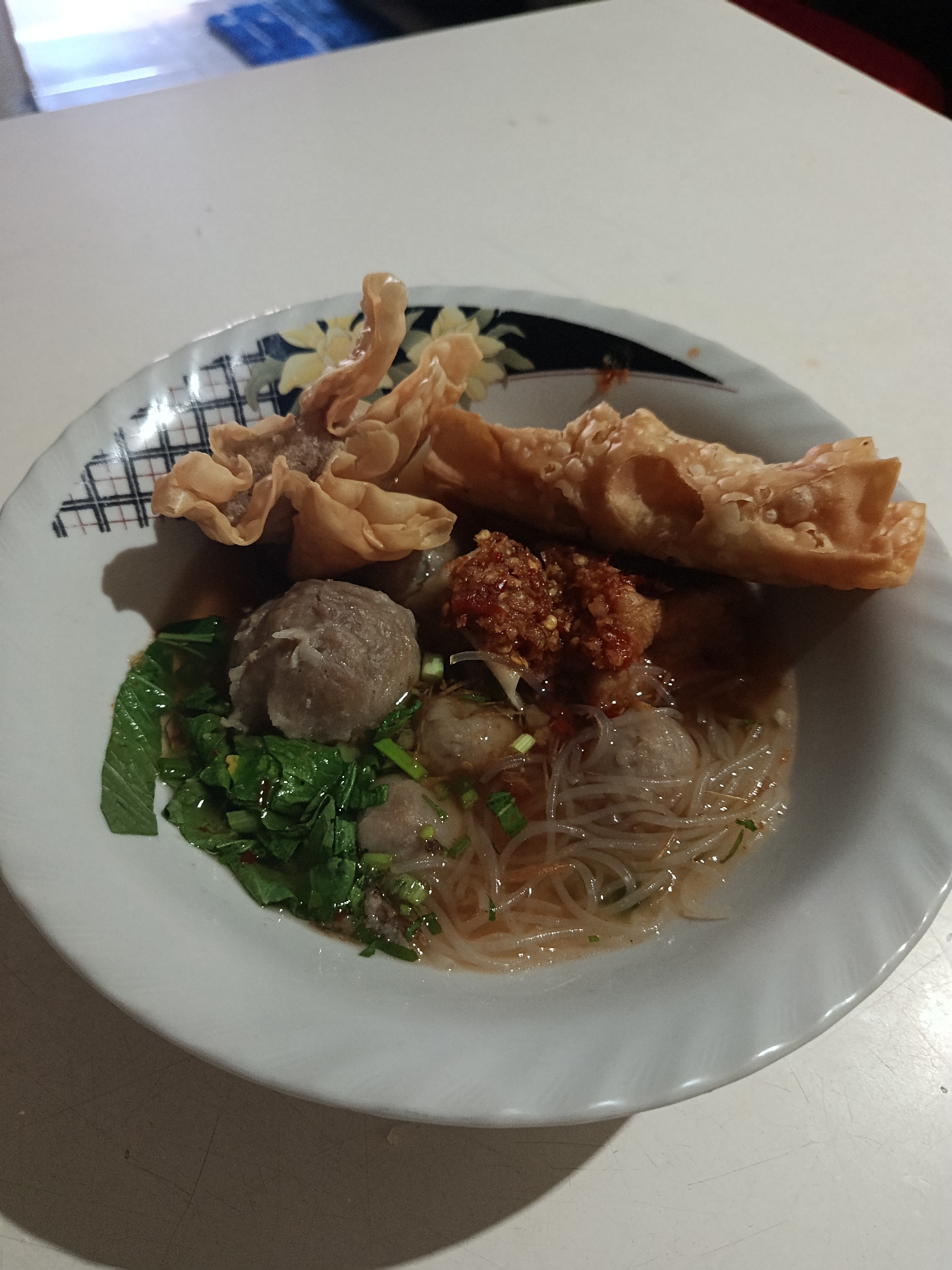 A high-angle shot shows a bowl of bakso in Keboananom, Indonesia. The bowl contains meatballs, noodles, greens, and fried wontons. The bowl sits on a white table.