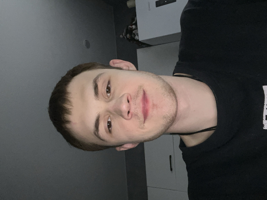 A young man with short, dark hair and light skin faces the camera with a slight smile. His eyes are brown, and he has a light beard. He wears a black t-shirt and a thin black string necklace. The background includes a gray wall and white cabinets. The image is slightly tilted, giving a sideways perspective.