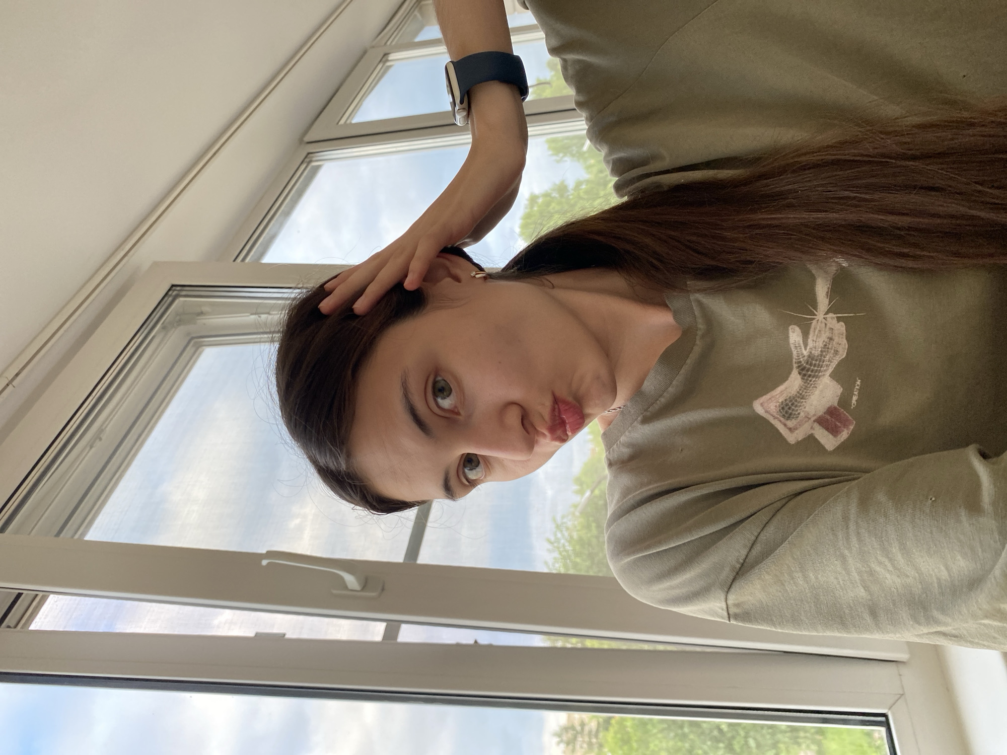 A young woman with long, dark brown hair pulled into a ponytail, who appears to be in her late teens or early twenties, is captured in a selfie taken indoors. She is wearing a loose-fitting, light olive green t-shirt with a graphic design on the front. Her left hand is raised and resting on her head, while her right arm is extended, wearing a dark blue smartwatch with a white face. She is making a pouting expression, with her lips pursed.
The background suggests an interior setting near a large window. Through the window, a cloudy sky and green trees are visible, indicating an outdoor environment. The window frame is white, and part of a white wall is visible above the window. The lighting suggests it is daytime. The overall scene is casual and personal.
Based on the provided information, it is not possible to determine the specific location in Votkinsk, Russia.