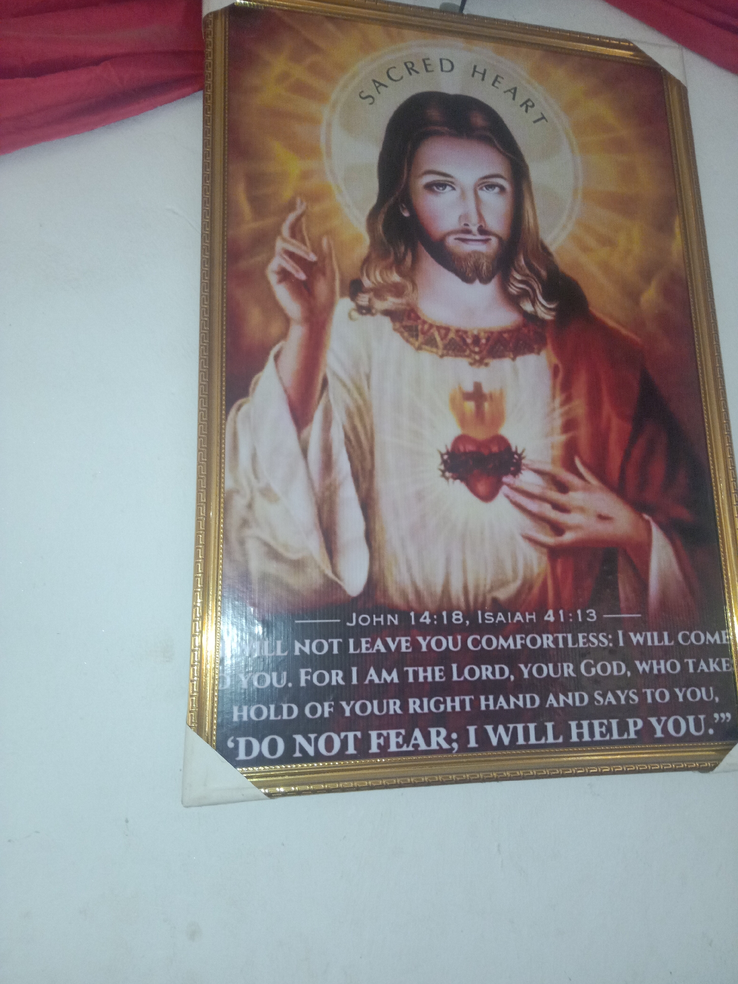 The image shows a framed artwork of the Sacred Heart of Jesus hanging on a white wall. The artwork features Jesus Christ with a halo and a radiant heart on his chest, surrounded by golden rays. Text is inscribed below the image, quoting scripture. A red fabric is draped above the artwork. The scene appears to be indoors. The specific location in Lokoja, Nigeria, is not identifiable from the visual information presented.