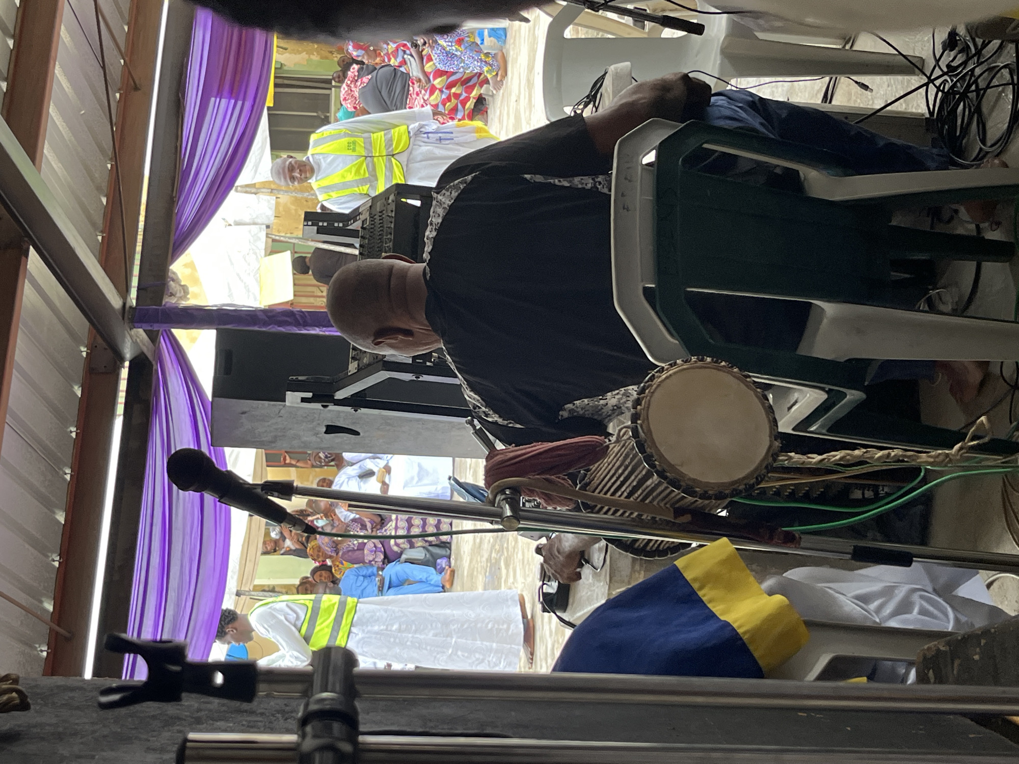 This media file captures an indoor gathering, possibly a celebration or a religious service, given the presence of people in traditional attire and a ceremonial drum. The scene is set under a metal roof structure, with purple and white drapes adding a decorative touch. In the foreground, a man in a black shirt is seated and appears to be playing a djembe drum. Another man in a reflective yellow vest and white clothing is visible in the background, suggesting a role in security or organization. Several other individuals, including women in colorful patterned clothing, are also present, indicating a social event.
The lighting suggests it's daytime. The overall mood seems to be one of engagement and community, with people gathered together. Some individuals are looking towards the front, where the music might be taking place, while others are engaged in conversation or observation. The image is oriented vertically, with the top of the scene appearing at the left. There is no visible text in the image. The presence of microphones and electronic equipment hints at a performance or a formal presentation component to the event.