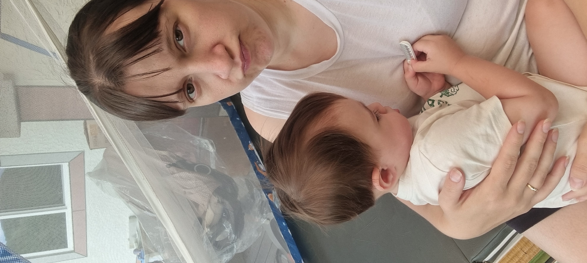 An indoor, eye-level shot captures a woman holding a baby in Dunnae-myeon, South Korea. The woman, positioned towards the top left of the frame, has dark hair with bangs framing her face. She looks directly at the camera with a neutral expression, wearing a simple white t-shirt. Her left hand, adorned with a thin gold ring on the ring finger, gently supports the baby.
The baby, with soft brown hair, is nestled against the woman, facing away from the camera, and appears to be sleeping or resting peacefully. The baby wears a light-colored t-shirt, possibly cream or light green, which features subtle patterns. One of the baby's small hands is holding a light-colored object, possibly a remote control or a small toy.
A light, translucent mesh fabric stretches across the foreground, partially obscuring the background but allowing elements to be seen through it. Behind this mesh, a light-colored wall with subtle geometric patterns is visible, along with what appears to be a window or a framed picture. Further details include a blue fabric with white patterns, possibly part of bedding or a playpen, and a dark gray mat or surface beneath the baby. The bottom right corner reveals a glimpse of a traditional tatami mat, reinforcing the East Asian setting. The overall scene conveys a calm and intimate moment between parent and child during what appears to be daytime. No discernible text is visible in the image.