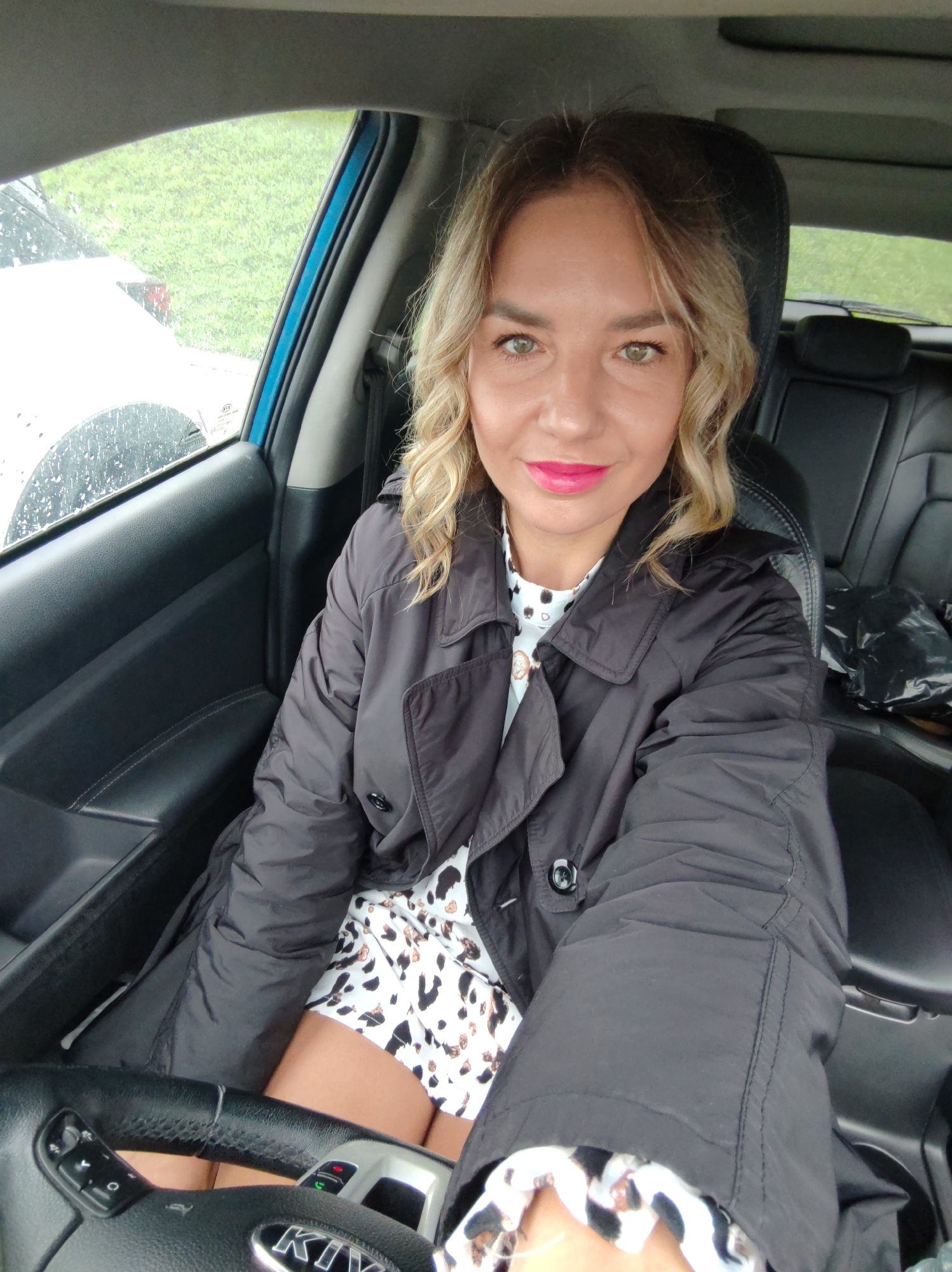 A woman is sitting in the driver's seat of a car. She is wearing a black coat over a patterned dress. Her hair is blonde and styled in waves. She has pink lipstick on and is looking directly at the camera. The car is parked, and the exterior is visible through the rain-streaked window. The interior of the car is black. The steering wheel has a KIA logo on it. Based on the limited environmental details visible through the window, it is not possible to confirm the location in the city of Petropavlovsk-Kamchatsky, Russia.