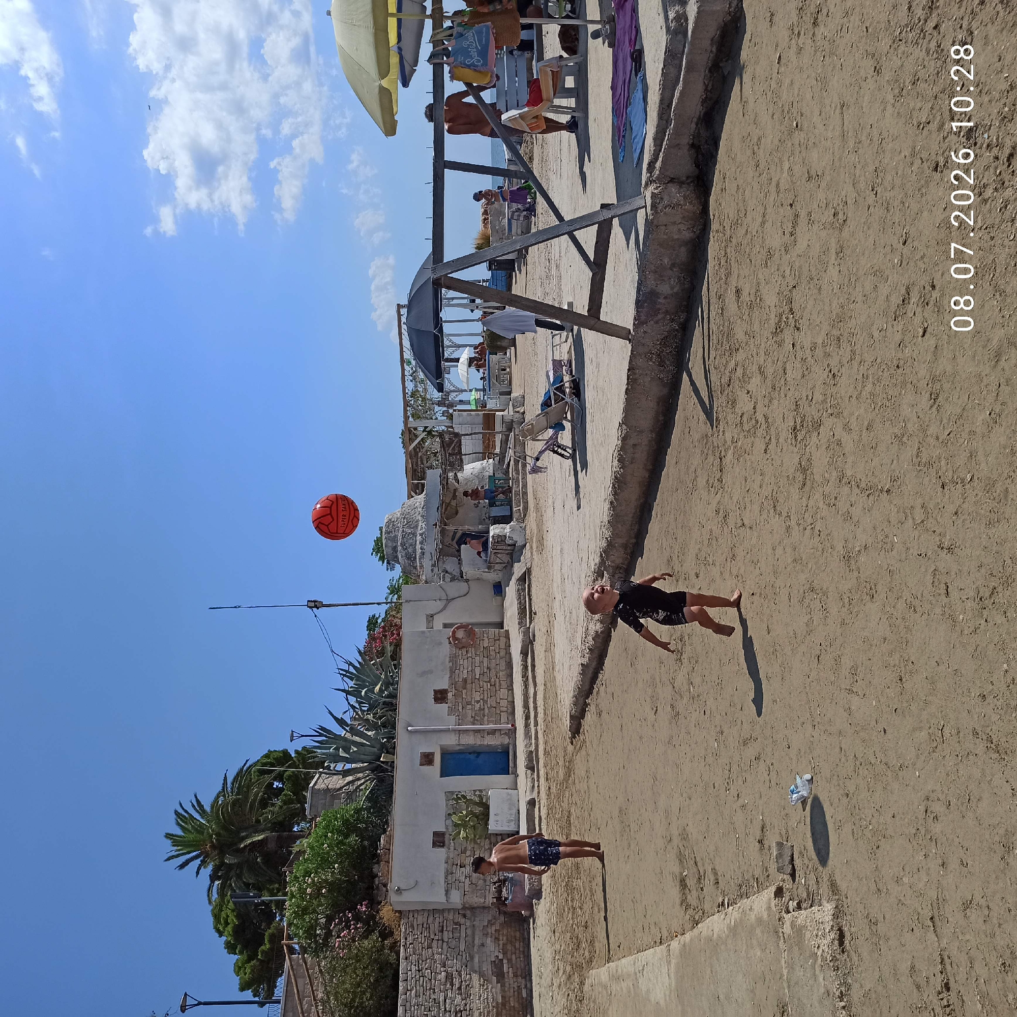 A group of people are on a sandy beach in Trani, Italy, on a sunny day. In the foreground, a young child in a black swimsuit is looking up at a red soccer ball in the air, with an older child standing to their left in blue shorts. Behind them, a building with a blue door is visible, as well as a swing set structure and beach umbrellas. In the background, more people are seen relaxing on lounge chairs and at tables. The sky is clear blue with a few scattered clouds.