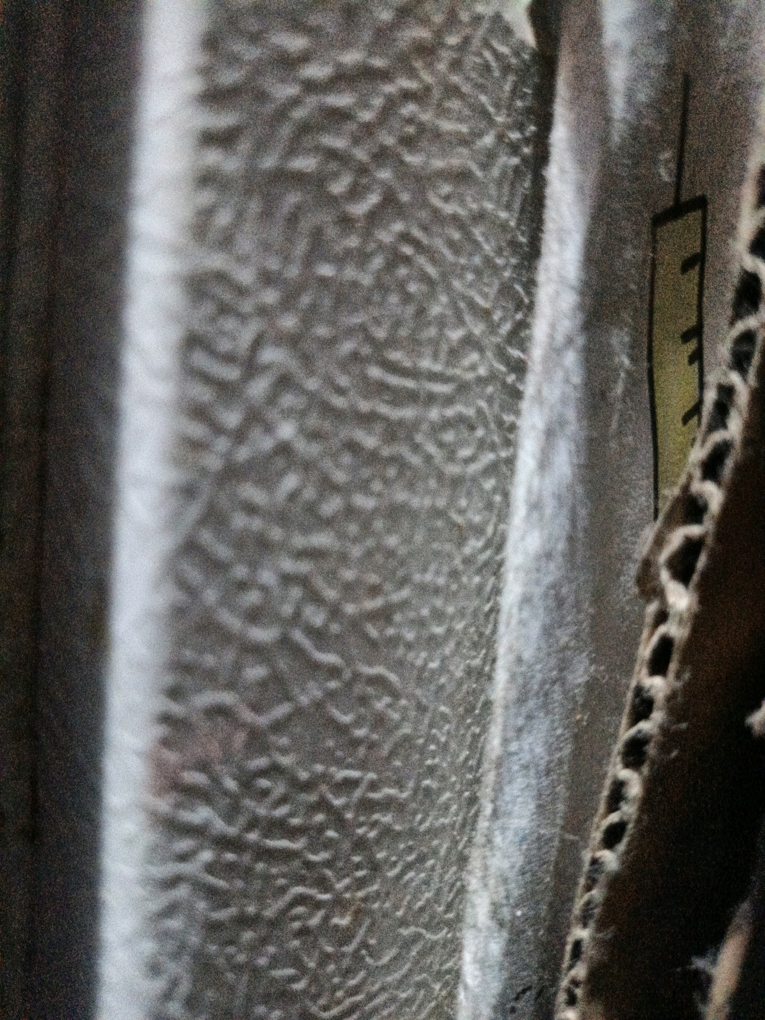 The image shows a close-up of a corrugated cardboard surface with a black and yellow label. The label appears to be a diagram or instruction, with a yellow rectangular area containing black lines and symbols. To the left of the cardboard, there is a blurred, vertical ribbed surface, possibly a wall or another material. The overall impression is one of a rough, industrial, or storage environment. The image does not contain any information that would allow for the determination of a specific location within West Jakarta, Indonesia.
