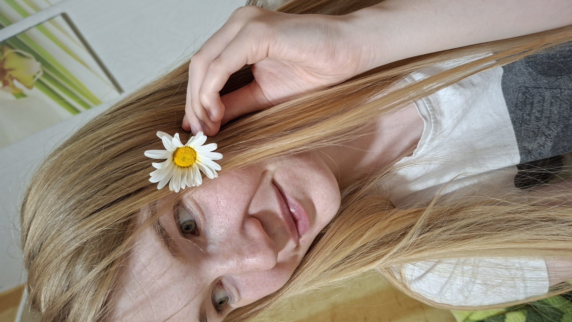 A young woman with long, blonde hair is positioned in the center of the frame. She has a daisy tucked behind her ear and is looking to her left with a slight smile. Her skin is fair and freckled. She is wearing a t-shirt with a white top half and a gray, patterned bottom half. Her left hand is visible, touching the flower. The background is blurred, but appears to be an indoor setting with a wall and some foliage visible. The image appears to be a close-up portrait. No specific location in Bolshoy Bukor, Russia can be determined from the image.