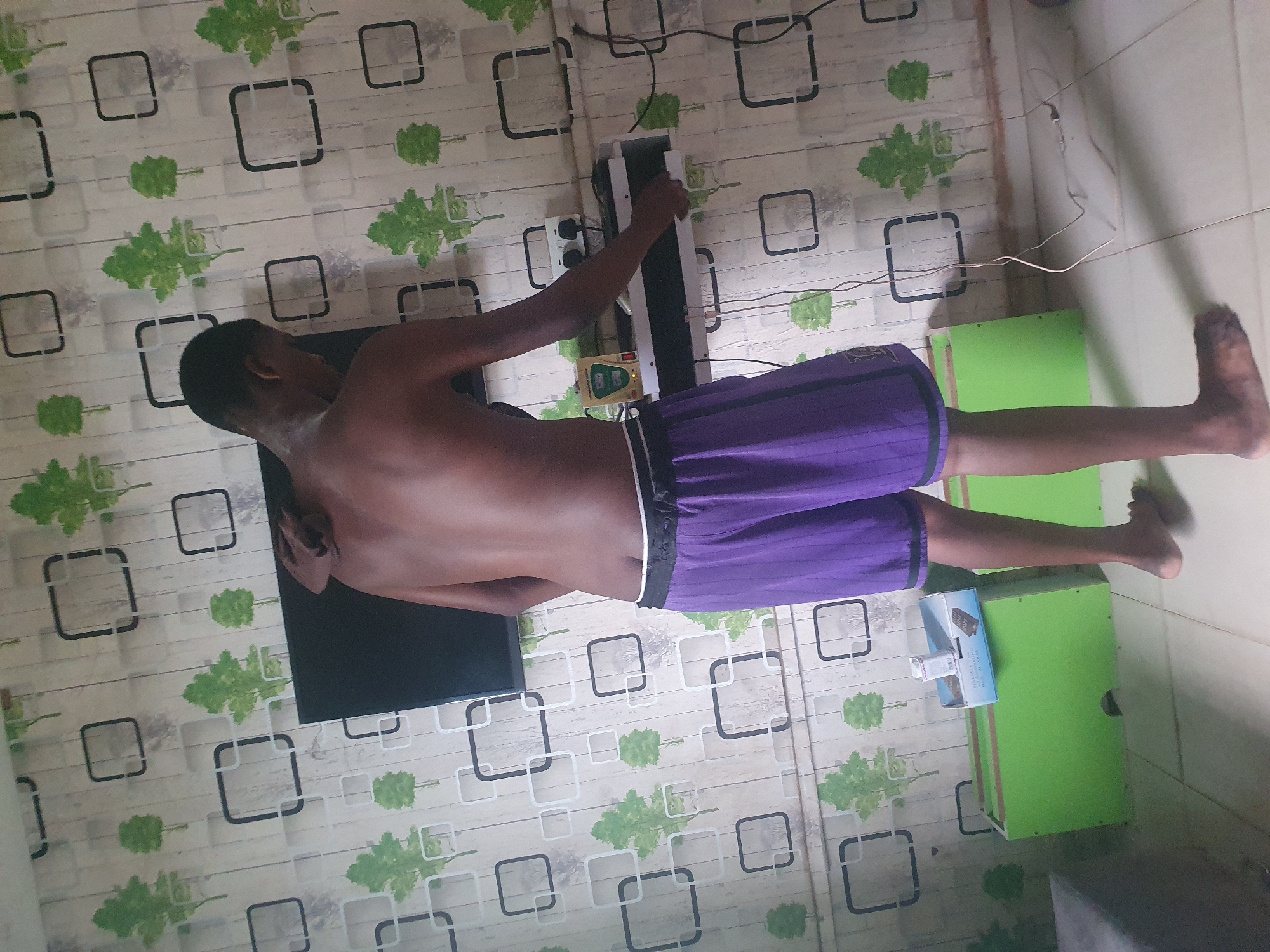 A shirtless individual wearing purple shorts stands in an indoor space with a distinctive wallpaper pattern of green foliage and square outlines, suggesting an interior room. The person is positioned next to a flat-screen television mounted on the wall, with electrical appliances and cords visible behind them. A green cabinet or box is visible to the right. The location is most likely a residential or commercial interior, but there is insufficient evidence in the image to confirm a specific city or location in Nigeria.