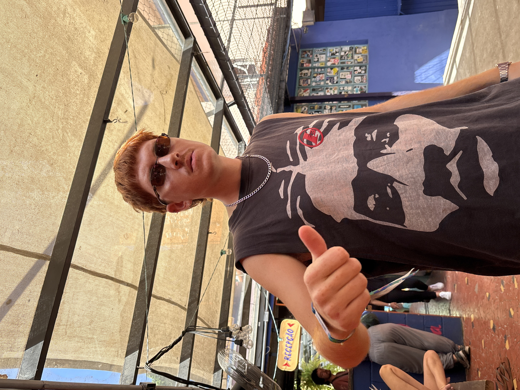 A young man with reddish hair and sunglasses gives a thumbs-up in an outdoor setting, likely in São Paulo, Brazil, based on the "Recepção" sign (Portuguese for reception). He wears a grey tank top with a large white and black print and a silver chain necklace. Behind him is a covered patio area with metal beams and a beige canopy. In the background, people are seated on blue mats, and a blue wall is covered with various stickers. A metal fence and building are visible further back. A fan and string lights hang from the patio structure. The ground is textured and painted in a reddish-brown hue.