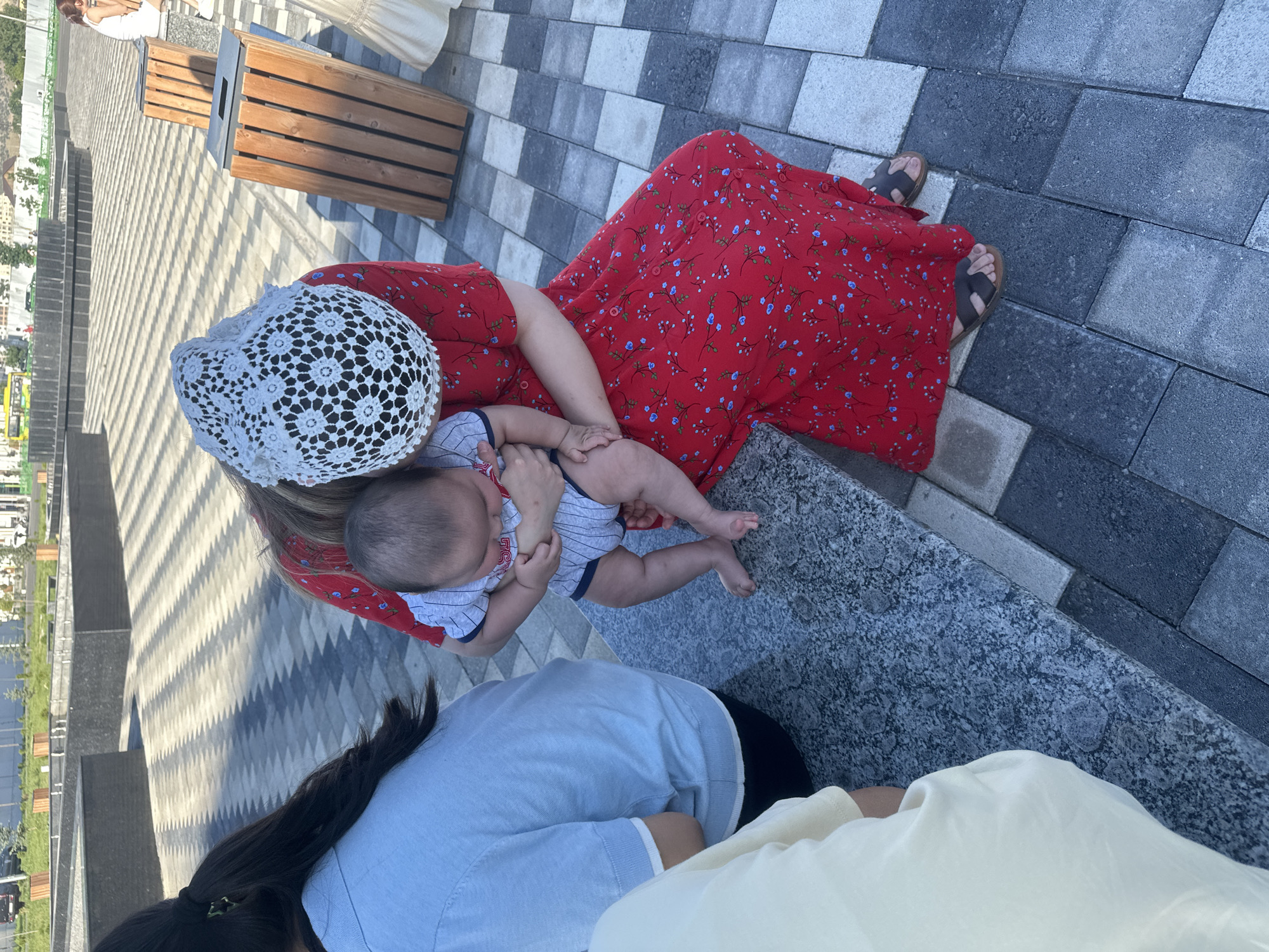 This media file captures a scene from above, showing several people sitting on a tiered stone and tile pathway. The primary focus is on a woman in a red floral dress, holding a baby who is wearing a striped onesie. The woman is also wearing a white lace head covering. To her left, another person in a light blue shirt is visible, and to the right, a glimpse of someone in a light yellow garment.
The setting appears to be an outdoor public space, possibly a park or a plaza, with elements like wooden benches and planters visible in the background, along with some greenery and buildings in the distance. The pathway is constructed with large, alternating grey and light-colored rectangular tiles, creating a geometric pattern. The lighting suggests it is daytime, likely with bright sunlight casting shadows. The overall mood seems relaxed and familial, with the woman cradling the baby. There is no visible text in the image. Based on the location context of Manas, Kyrgyzstan, this could be a scene from a local gathering or outing.