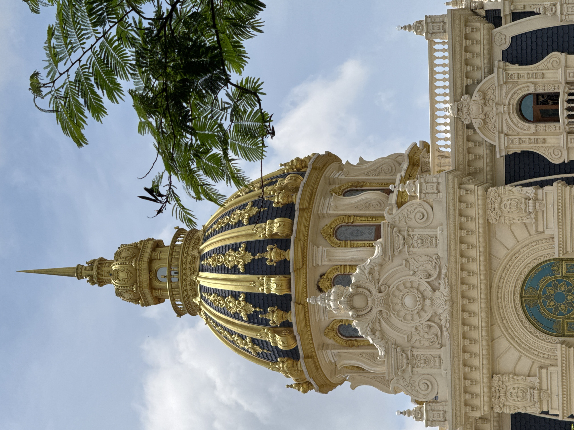This image captures a close-up view of a prominent golden dome and a section of a building's facade, set against a partly cloudy sky. The dome is intricately designed with ornate golden patterns against a dark blue tiled surface, topped with a sharp spire. The building itself is adorned with elaborate cream-colored decorative plasterwork, featuring scrollwork, molding, and architectural details around windows and along the roofline. A balustrade with decorative posts runs along a section of the building. Lush green foliage from a tree extends into the frame from the upper right, partially obscuring the sky and the top of the dome. The architectural style suggests a grand, possibly historical or themed structure. This building is the Dinh I Bao Dai (Bao Dai's Summer Palace) in Da Lat, Vietnam. There are no people or discernible actions depicted in the scene.